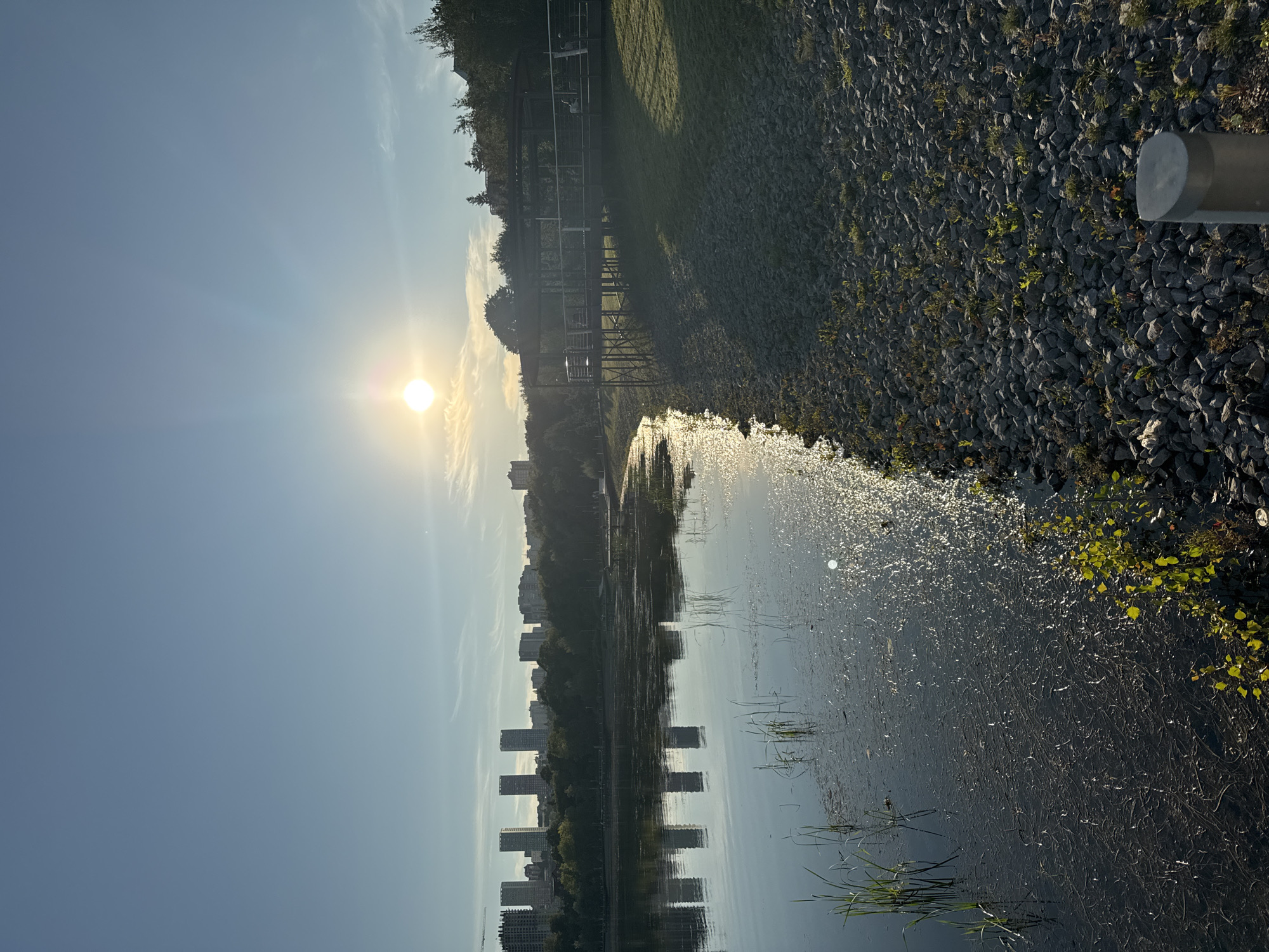 This is a landscape photograph taken at sunrise or sunset in Ufa, Russia. The scene depicts a tranquil body of water with reflections of distant city buildings and trees. The foreground features the rocky shore of the water body, with patches of grass and small plants growing between the stones. On the right side of the image, there is a wooden pier or boardwalk structure with a roof, supported by metal stilts. The sky is clear blue with a bright sun low on the horizon, casting a warm glow across the scene and creating shimmering reflections on the water. The cityscape in the background consists of several tall, modern buildings lined up against a backdrop of lush green trees. The overall atmosphere is peaceful and serene, showcasing a natural environment integrated with urban development.