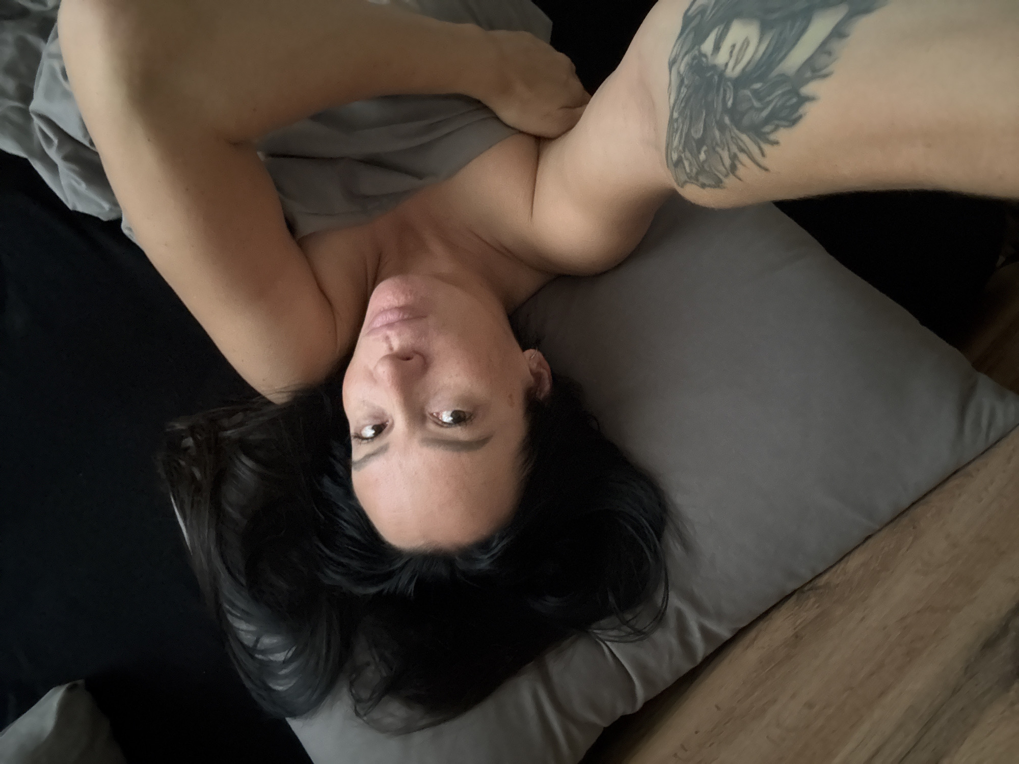 A woman with long, dark hair is lying in bed, looking at the camera. She is partially covered by a gray blanket. She has a tattoo on her left arm. The bed appears to be in a bedroom. The scene does not provide any specific details that would allow for determination of its location in Chaikovsky, Russia.