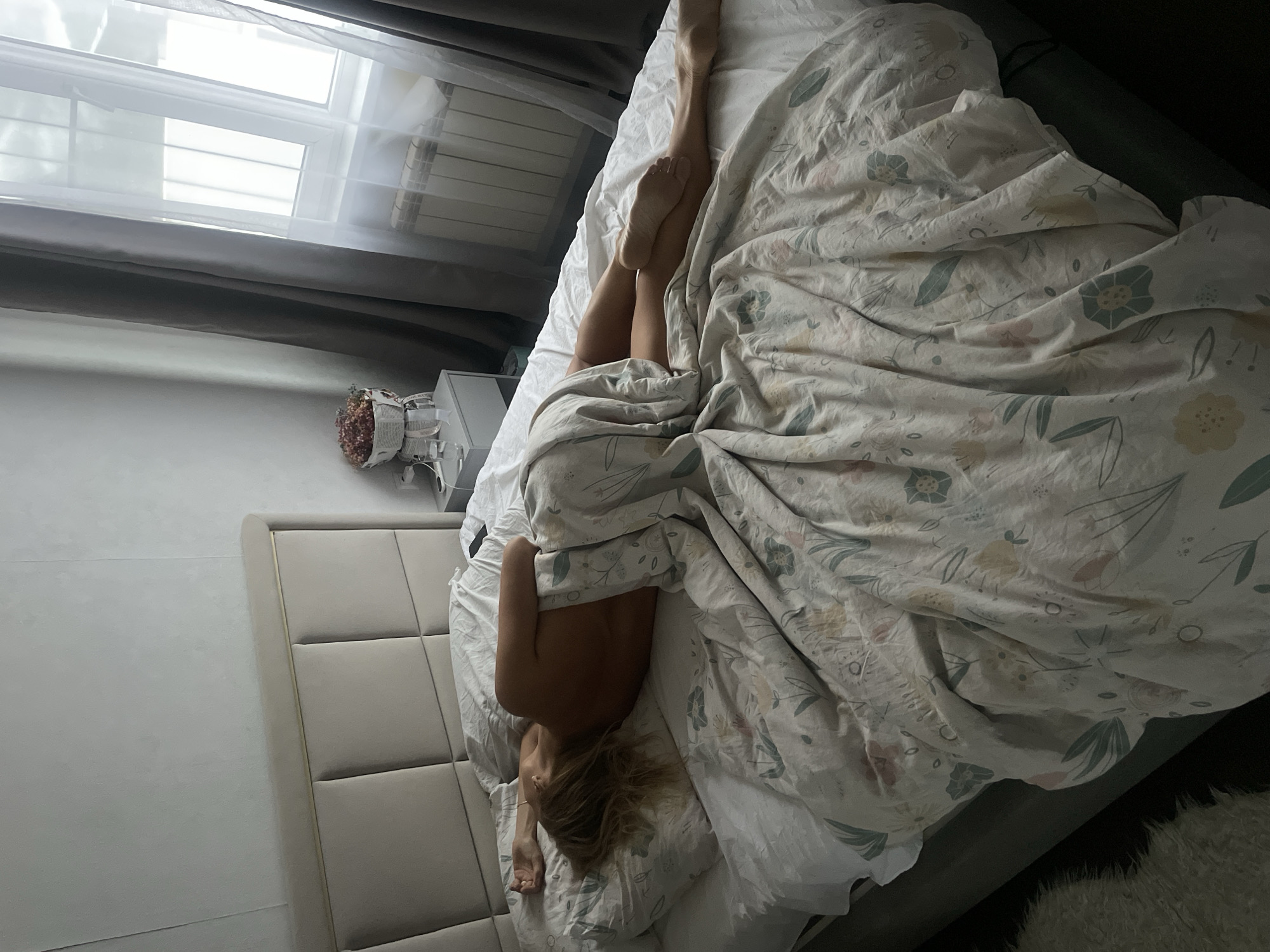 A high-angle shot depicts a woman lying in a bed covered with a floral-patterned duvet. Her legs are extended upwards, while her torso and head are partially covered by the duvet. A headboard is visible on the left side of the bed. Natural light streams in from a window in the background. To the left of the bed, there is a bedside table with a flower arrangement on top. The location is not identifiable as the city of Baranovka, Russia, from this image alone.