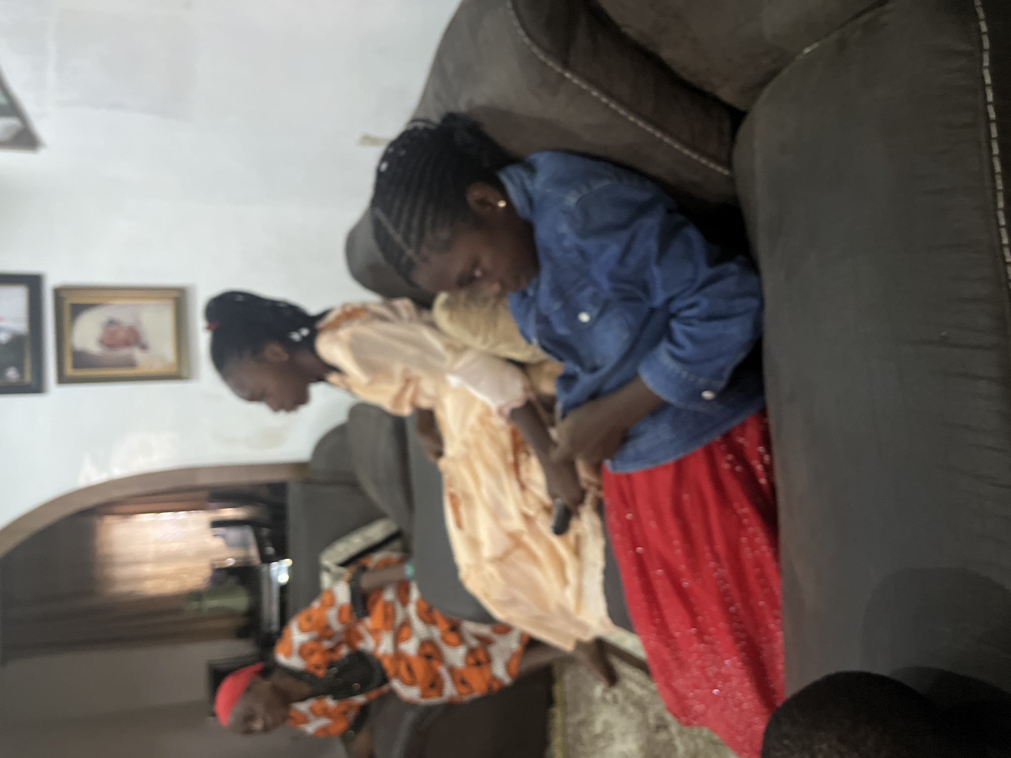 In Jos, Nigeria, three individuals are seated on a dark gray sectional sofa in a living room. On the left, an older woman wearing a red headscarf and an orange and white patterned traditional outfit is partially visible, facing to the left. In the center, a young girl with braided hair and a light peach-colored dress is looking to the right. To the right, another young girl with braided hair, wearing a blue denim jacket and a red sequined skirt, is looking down at a remote control in her hands. The room is dimly lit, with framed pictures on the wall in the background. A large archway is visible to the left of the sofa, leading to another part of the room.