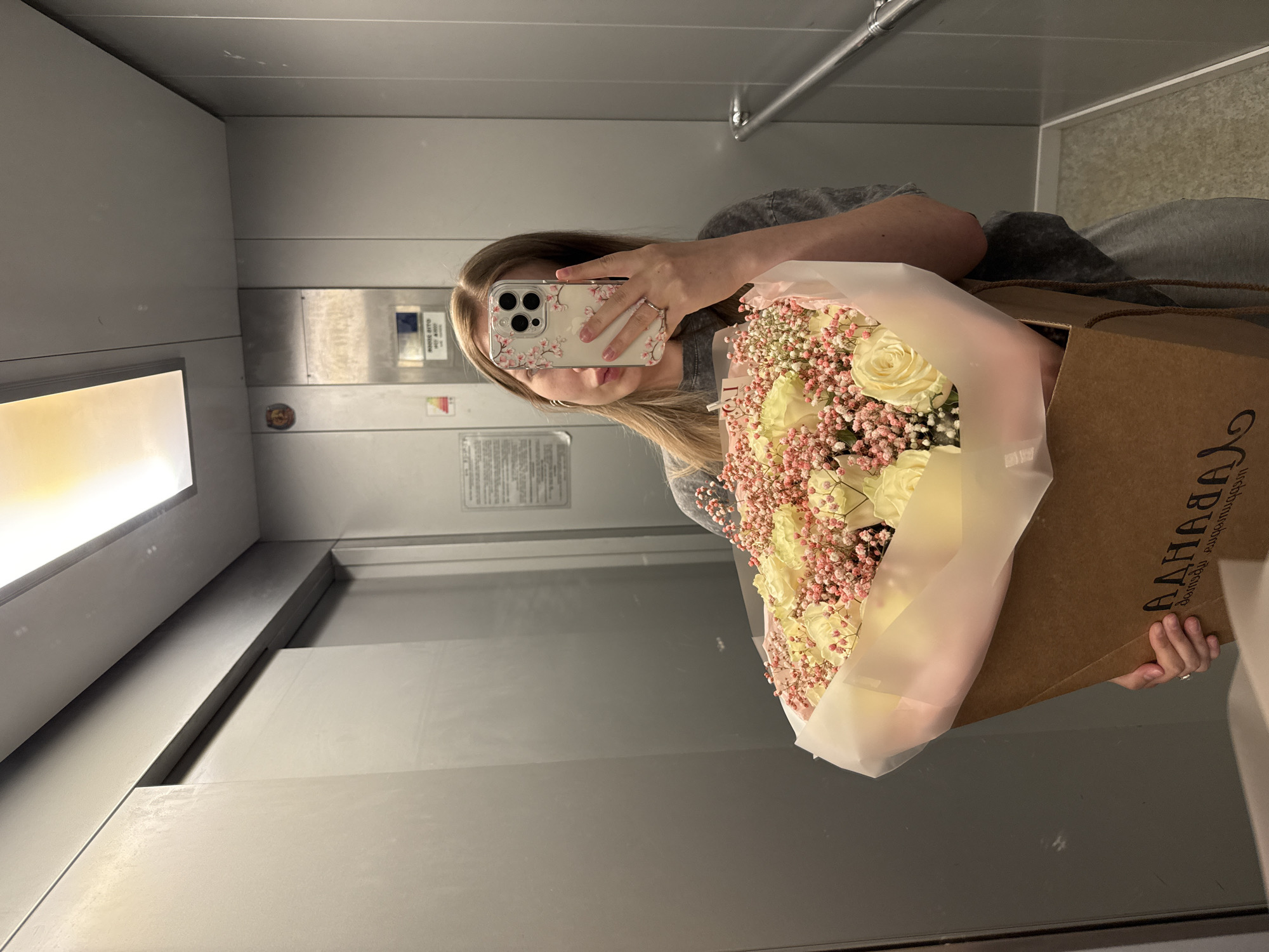 A young woman with blonde hair and light-colored eyes is taking a selfie in an elevator. She is holding a large bouquet of white roses and pink baby's breath in a brown paper box. Her phone, with a floral case, is held up to take the picture. The elevator walls are silver, and there is a metallic handrail on the right side. The floor of the elevator is visible, with a patterned tile design. There is a light fixture on the ceiling, and a small sign on the back wall. The logo on the flower box reads "ADANAHA HOUSE". The scene appears to be in an apartment building or a hotel in Chaikovsky, Russia.