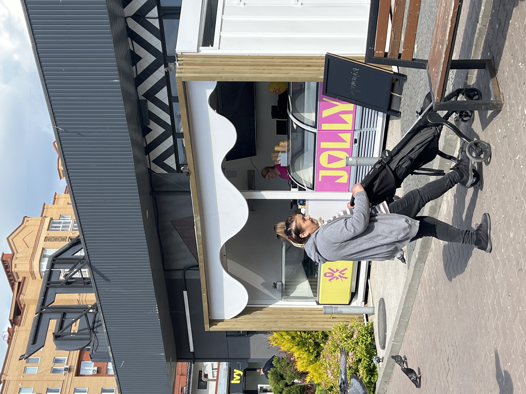 A woman pushes a stroller past a food stand with the name "JOLLY" in large pink letters. The stand is white with wood trim and has a striped awning. Inside, a person wearing a pink shirt is visible behind a glass display. A blackboard with handwritten text is propped up next to the stand. The woman is wearing a grey hoodie and black pants. The scene takes place outdoors on a paved sidewalk, with a building and trees in the background. The lighting suggests it is daytime. There are several pigeons on the ground near the stroller and the sidewalk. The context provided indicates the location is Ternopil, Ukraine.