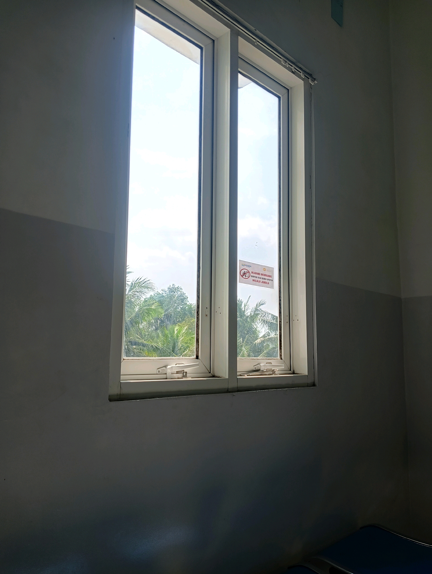 A tall, narrow window with two sashes is centered in the frame, looking out onto lush green trees and a bright sky with scattered clouds. The window frame is white, and a red and white sticker is affixed to the left sash. The interior walls are painted in a gradient of gray, with a darker shade towards the bottom of the frame and a lighter shade towards the top. The lighting suggests it is daytime, with sunlight streaming through the window and casting shadows on the walls. In the bottom right corner of the image, a portion of what appears to be blue seating is visible, indicating the interior is likely a waiting area or a room in a building. The location is specified as Purwareja, Indonesia.