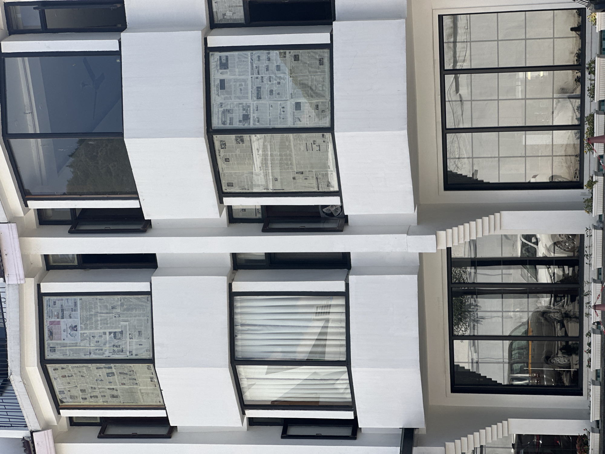 The image shows the exterior of a multi-story building, likely a hotel or apartment complex, with a modern architectural style. The building is painted white and features large, dark-framed windows. Some windows are covered with newspapers from the inside, presumably for privacy or insulation. The upper floors have bay windows, while the ground floor has large, floor-to-ceiling windows that reflect the surroundings. Parked cars are visible in the reflection of the ground floor windows. Planters with small plants are situated along the base of the building. The overall scene suggests an urban setting with a touch of residential or commercial activity. This is a generic building facade that could be found in many cities; there are no specific visual cues that definitively place it in Srinagar, India.