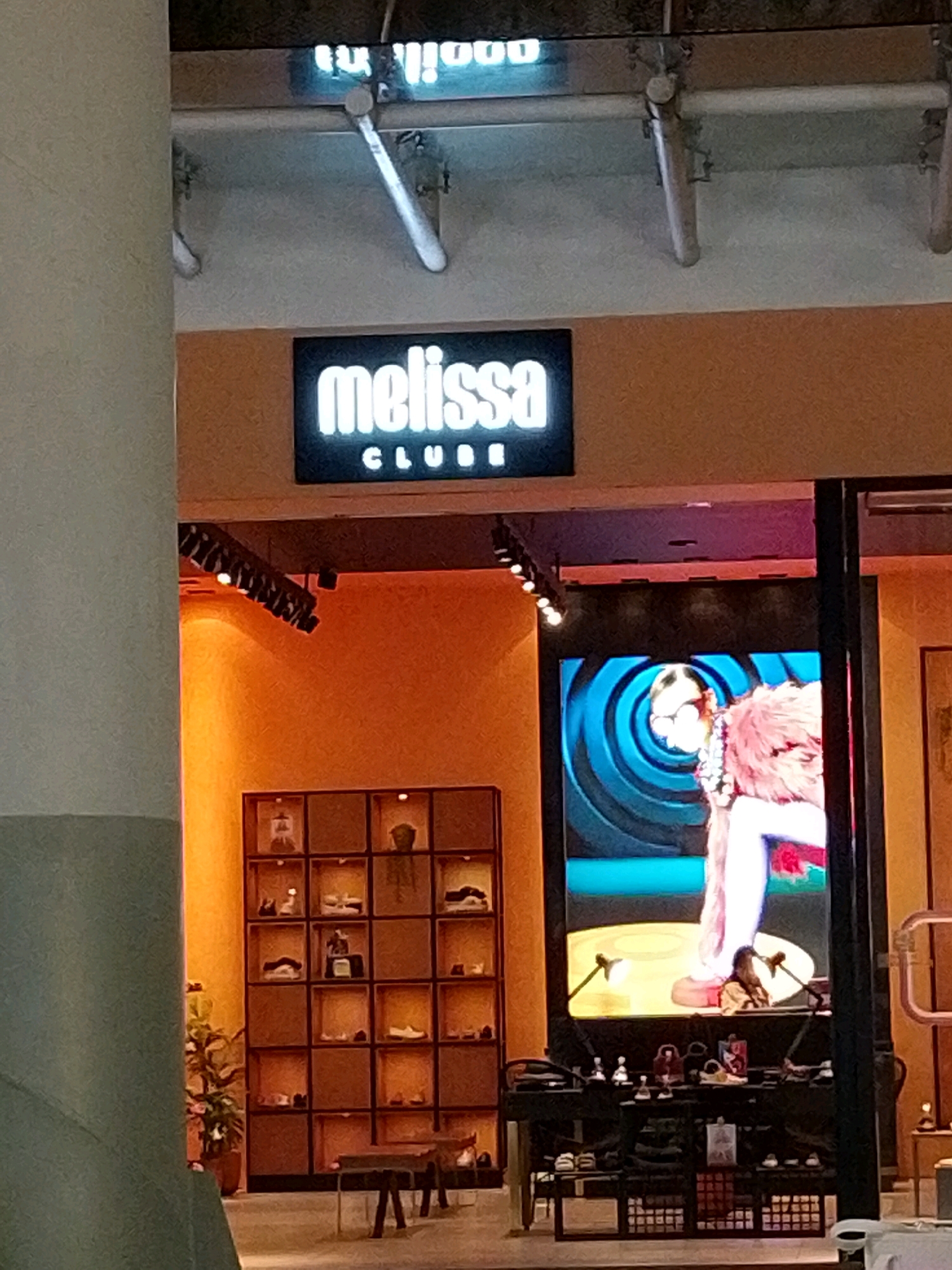 The image shows a storefront with a neon sign that reads "melissa CLUB." Below the sign, there is a large digital display screen showing a person with glasses and a pink fluffy garment. To the left of the display screen, there is a shelf unit filled with shoes. The storefront appears to be located in a mall or shopping center. The specific location is unclear from the image alone, but the presence of a "Melissa" store suggests it could be in a city with a presence of this shoe brand.