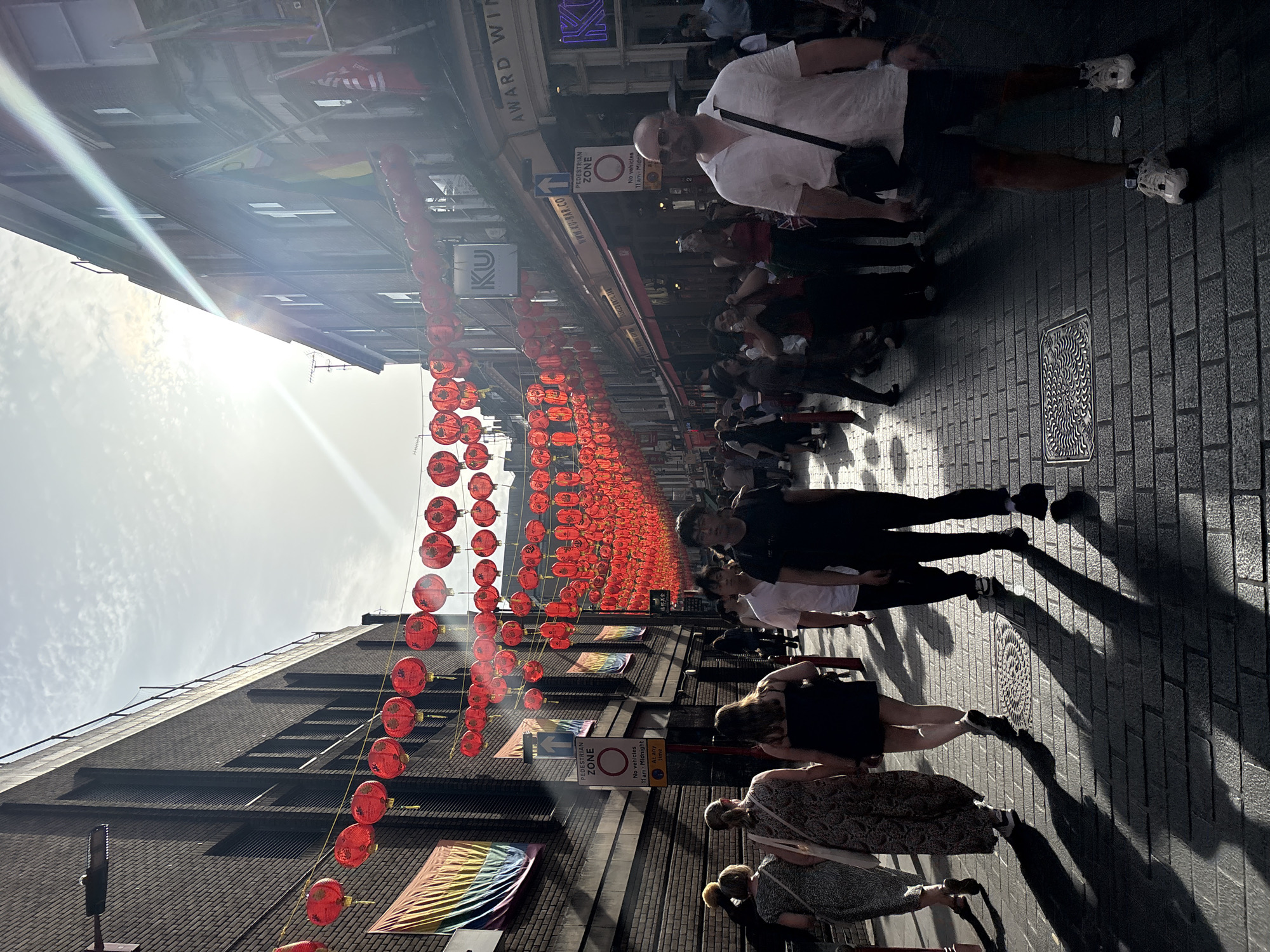 The image shows a street scene in London's Chinatown, United Kingdom, characterized by a street festooned with numerous red Chinese lanterns strung overhead. A few pedestrians stroll along the brick-paved street, their shadows elongated due to the sun's position. Various shops and buildings line the sides of the street. Signs indicating a pedestrian zone and bar details are visible. Some rainbow flags, symbols of LGBTQ pride, adorn the buildings.