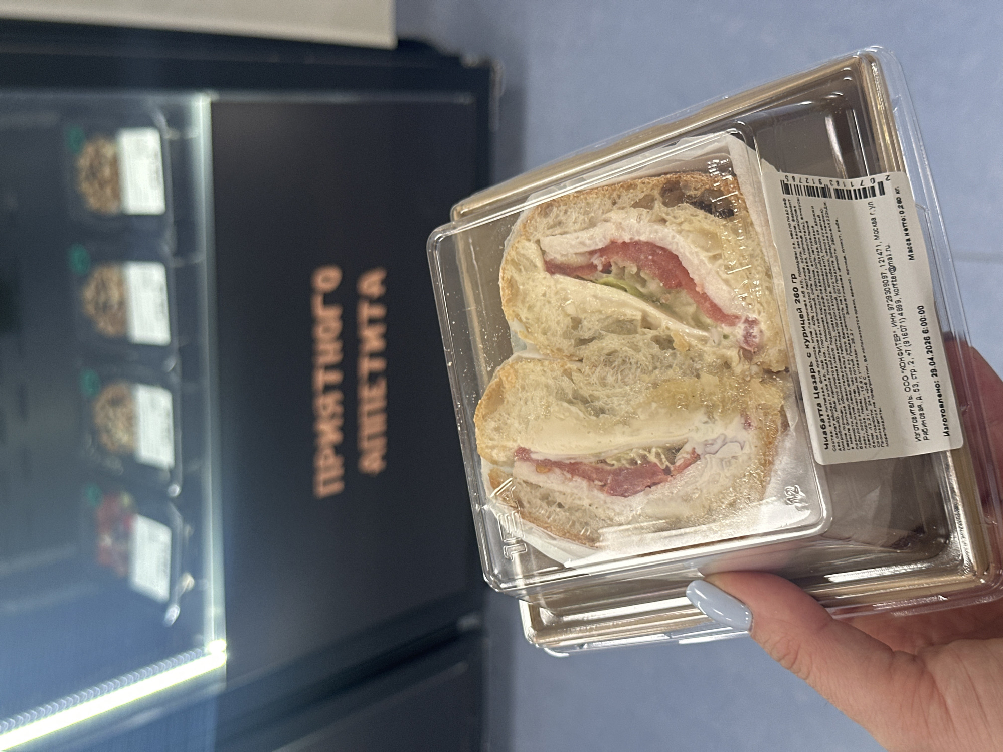 A close-up, slightly tilted shot shows a person's right hand with light blue nail polish holding a clear plastic container with a sandwich. The sandwich, labeled "Чиабатта Цезарь с курицей" (Chiabatta Caesar with chicken), features light-colored bread, chicken slices, a reddish ingredient (possibly cured meat or tomato), and white sauce. The container's label, prominently displayed, provides detailed information in Russian, including ingredients, nutritional values (200 kcal/822 kJ, 12.2g protein, 15.9g fat, 26.2g carbohydrates), storage conditions (24 hours at +2 to +6 °C), allergen warnings for nuts, peanuts, mustard, sesame, fish, and seafood.
The manufacturer is identified as OOO "KONFITER" located in Moscow, Ryabinovaya street, d. 53, building 2. The manufacturing date is stated as 29.04.2026 6:00:00, with a net weight of 0.200 kg. A barcode is also visible.
In the blurred background, a dark-colored vending machine or refrigerated display case is visible, containing other packaged food items. The machine displays the phrase "ПРИЯТНОГО АППЕТИТА" (Enjoy your meal or Bon appétit) in orange letters. The scene suggests an indoor setting, likely a commercial or institutional space in Khimki, Russia, where the sandwich was either purchased or is about to be consumed.