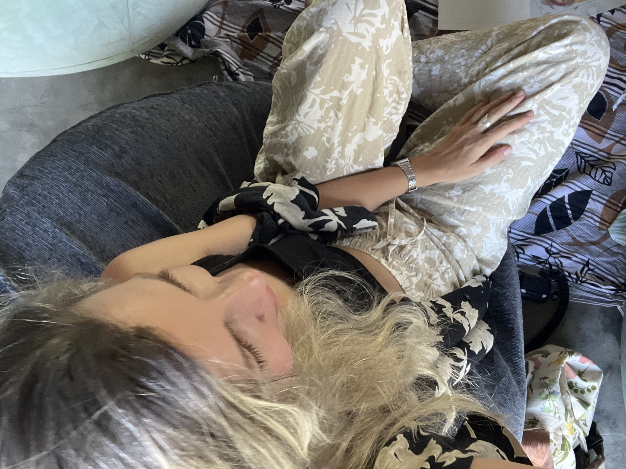 This is a high-angle, close-up shot of a person lying down, with their eyes closed. The subject has blonde hair and is wearing patterned clothing. Their left hand rests on their lap, adorned with a silver ring. A watch is visible on their left wrist. The setting appears to be indoors, with various patterned fabrics and a dark-colored chair or cushion visible. The overall mood is relaxed and peaceful, suggesting a moment of rest or sleep. The lighting is natural, indicating it could be daytime. The provided location context is Weligama, Sri Lanka. There is no visible text in the image.