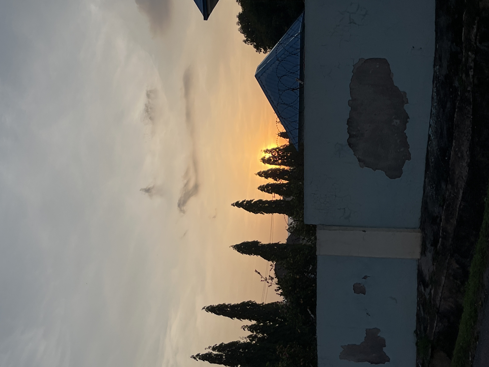 The image depicts a sunset scene with a sky transitioning from pale blue to a warm orange glow as the sun dips below the horizon. Silhouetted against the vibrant sky are tall, slender trees that line a fence. The fence itself is a light blue color and shows signs of peeling paint, revealing patches of a darker surface underneath. A section of the fence is a lighter, yellowish color, possibly a pillar or a different material. Beyond the fence and trees, a blue, pyramid-shaped roof structure is visible, topped with barbed wire. The overall environment suggests an outdoor setting, possibly a residential or semi-commercial area, with a focus on the natural light of the sunset and man-made structures. The scene is devoid of any visible people or animals. There is no indication in the image that places it specifically in Abuja, Nigeria.