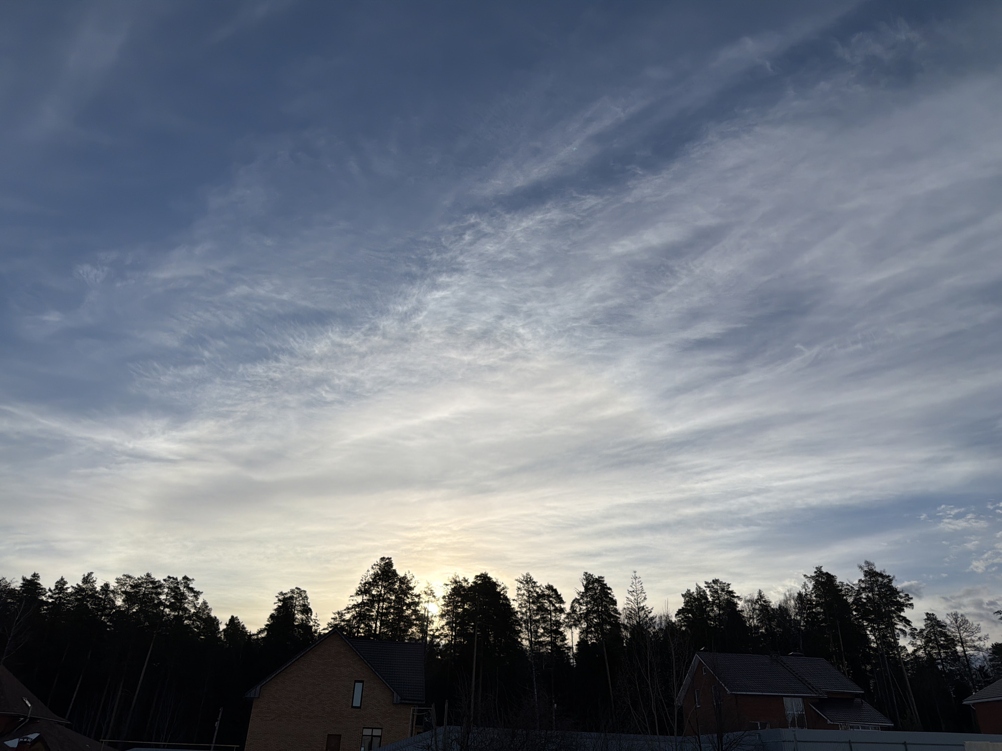 This image depicts an exterior scene in a residential area of Chaikovsky, Russia, at what appears to be either sunrise or sunset. The sky, which occupies approximately two-thirds of the frame, is largely blue with extensive, wispy cirrus clouds distributed throughout. A distinct bright yellow-white glow is visible on the horizon, slightly left of center, indicating the low position of the sun and backlighting the scene.
A dense, dark silhouette of a forest, composed primarily of tall evergreen trees (likely pines) and some bare deciduous trees, stretches across the middle ground. In front of this tree line, three residential structures are visible in the lower third of the image. On the far left, a portion of a brick house with a dark-tiled roof is visible. To its right, a two-story brick house with a dark-tiled roof features multiple windows and what appears to be construction scaffolding or a similar structure immediately adjacent to its right side. A small section of a third house with a dark roof is visible on the far right edge of the frame. The immediate foreground is partially obscured by a low, light-colored fence or barrier in front of the central-right house, with the ground appearing to have a thin, shadowed covering, possibly snow or frost. No individuals or specific actions are depicted.