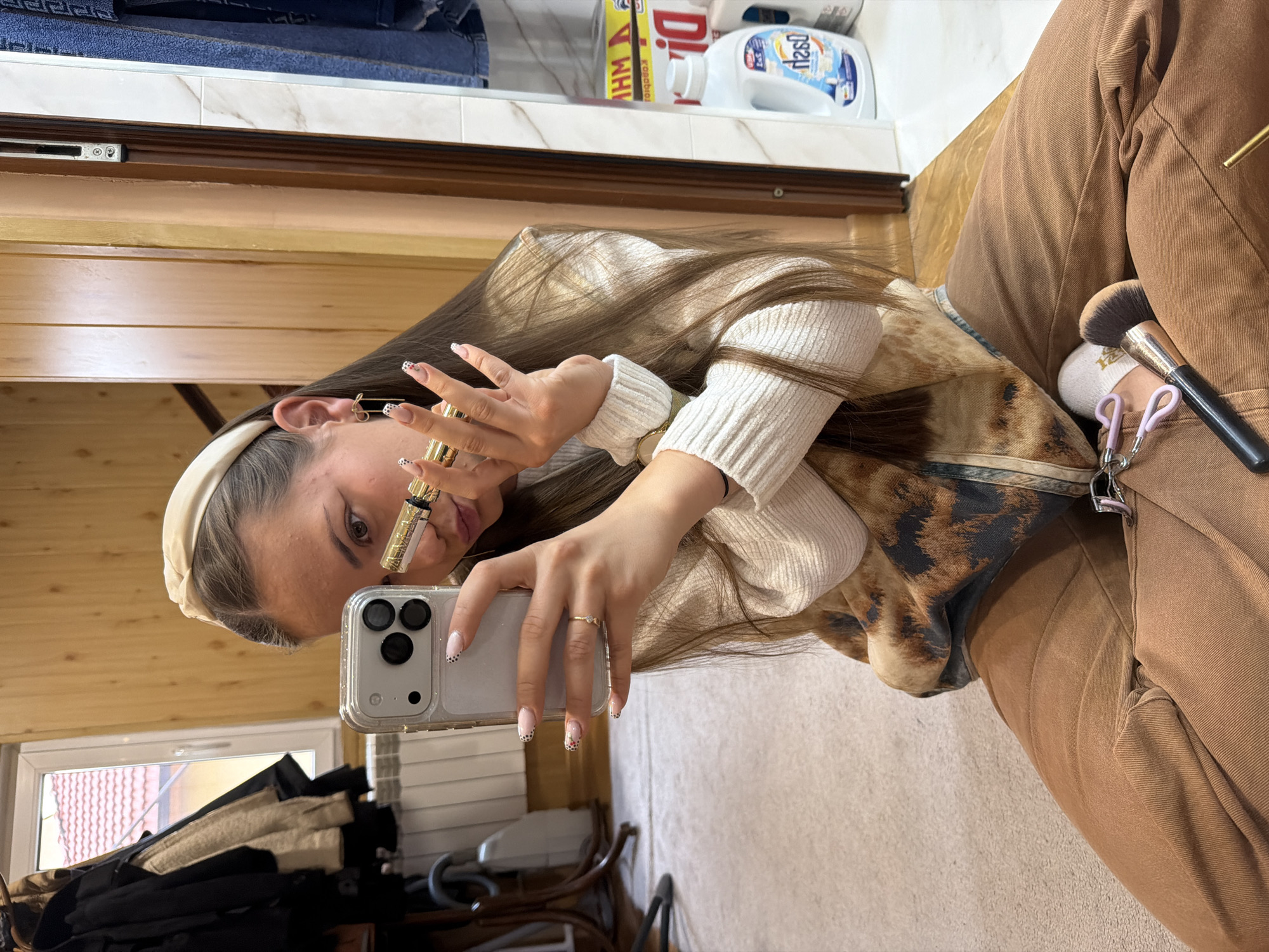 The image depicts a person taking a selfie indoors. The individual is light-skinned with long brown hair, wearing a cream-colored headband and a light-colored sweater with a brown and black patterned vest. They are holding a smartphone in one hand and a small, gold-colored cosmetic tube in the other, appearing to apply it. The phone has a clear case with black polka dots on the nails holding it.
A makeup brush and eyelash curler are visible on their lap, which is covered in brown pants. The background includes a wooden wall and a glimpse of a shelf with household cleaning products. A window is also visible, hinting at an exterior view.
There is no definitive evidence in the image itself to confirm the location is Buhovo, Bulgaria.