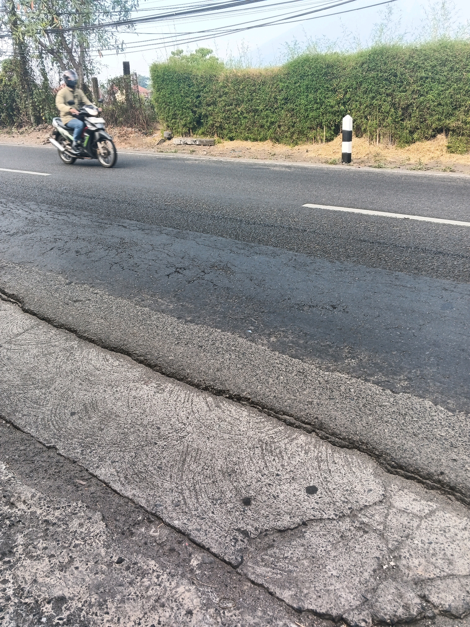 A person rides a motorcycle on a cracked asphalt road in Ungaran, Indonesia. The person is wearing a light brown jacket, blue jeans, and a dark helmet. The motorcycle is dark colored with silver accents. The road is lined with a dense green hedge on the right side of the image. A black and white striped post stands next to the hedge. Overhead power lines are visible against a clear sky. The foreground of the image shows the texture and cracks of the road surface.