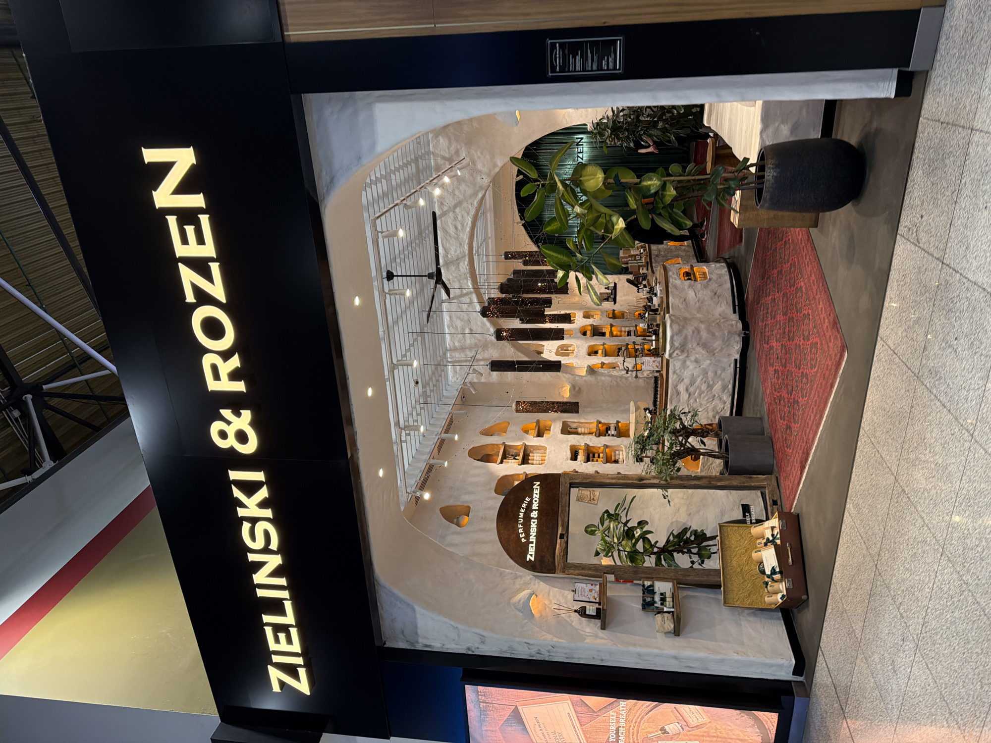 The image shows the exterior of a perfume shop called ZIELINSKI & ROZEN. The shop has a distinctive arched entrance with a rustic, white, textured facade. Inside, there are shelves stocked with various products, decorative lighting fixtures hanging from the ceiling, and plants. A large mirror is visible near the entrance, framed by wood. A red patterned rug is placed in front of the shop, and a suitcase filled with bottles is displayed on the floor to the left. The shop's name is illuminated in gold lettering on a black sign above the entrance. The surrounding environment appears to be a commercial area, possibly an airport or shopping mall, with tiled flooring and other store fronts visible in the background. No people are clearly identifiable in the foreground, though figures can be seen inside the shop in the background. The location is identified as Perepechino, Russia.