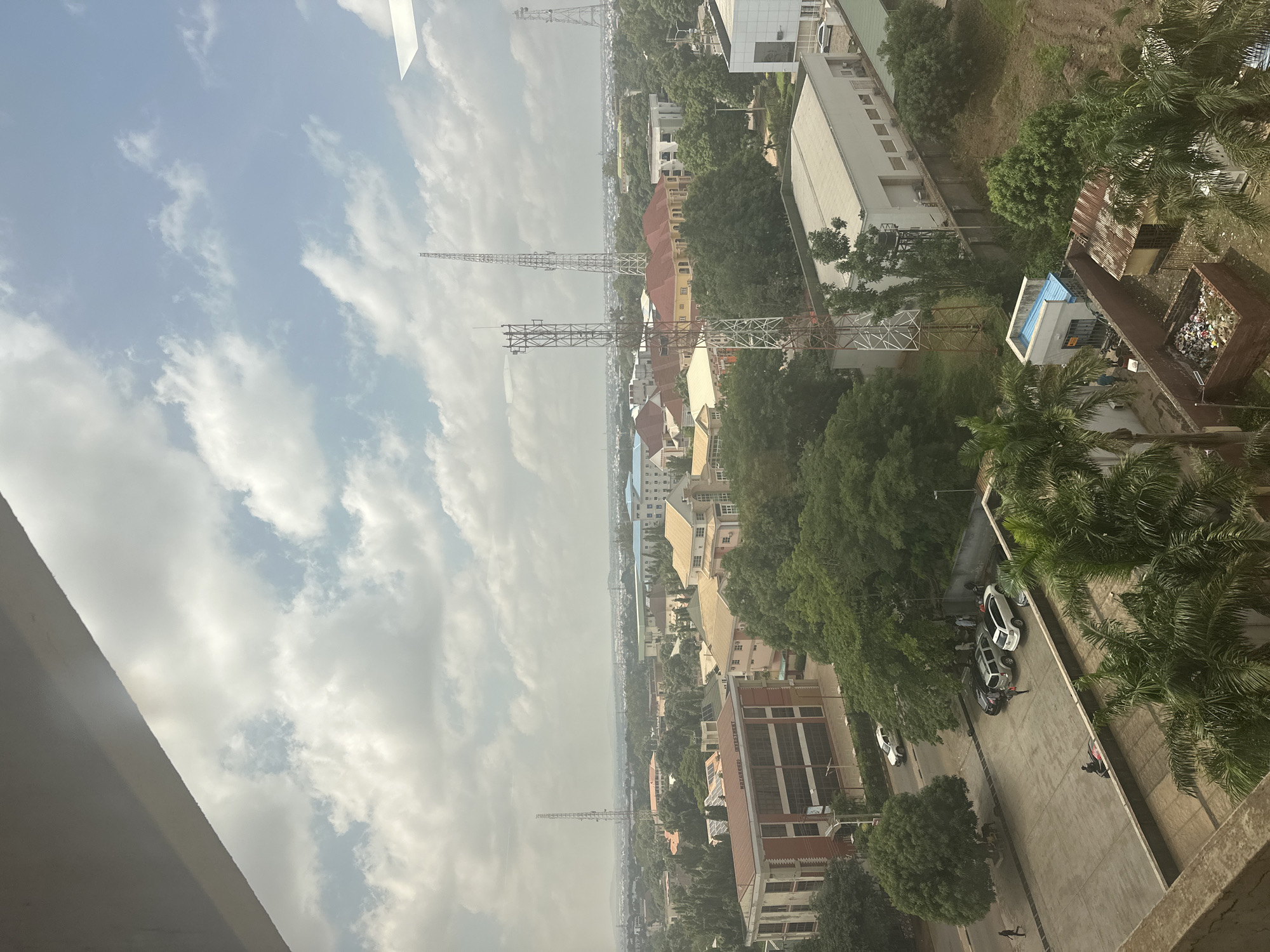 This is a high-angle, wide shot of a cityscape, likely Unguwar Sarki, Nigeria, on a cloudy day. The foreground is dominated by the edge of a concrete structure, possibly a balcony or rooftop, casting a shadow that partially obscures the view. Below, a paved area with several cars and pedestrians is visible. Among the vehicles, a white SUV and a white sedan are parked, while a dark SUV is also present. Two individuals are seen walking on the paved area, one near the cars and another further away. A person is also visible on a motorcycle.
Beyond this immediate area, the scene opens up to a dense urban landscape characterized by numerous trees and buildings. Several communication towers, including tall, lattice-style structures, dot the skyline, indicating a functional telecommunications hub. Buildings range in architectural style, with some featuring red brick facades and others appearing to be modern office or residential blocks with light-colored exteriors and tiled roofs in shades of orange and pink.
Greenery is abundant, with mature trees interspersed throughout the urban environment, suggesting a well-established and possibly well-maintained area. In the mid-ground, a long, rectangular building with a light-colored, flat roof is prominent, and further beyond, a sprawling city extends towards the horizon, where a hazy skyline suggests a larger metropolitan area.
To the right of the image, closer to the foreground, there is a small, makeshift structure with a corrugated metal roof, adjacent to a brick enclosure containing a substantial amount of refuse. This area contrasts with the more organized appearance of the buildings and infrastructure. The overall impression is one of an active, developing urban environment with a mix of residential, commercial, and infrastructural elements, set against a backdrop of lush vegetation and a somewhat overcast sky.
