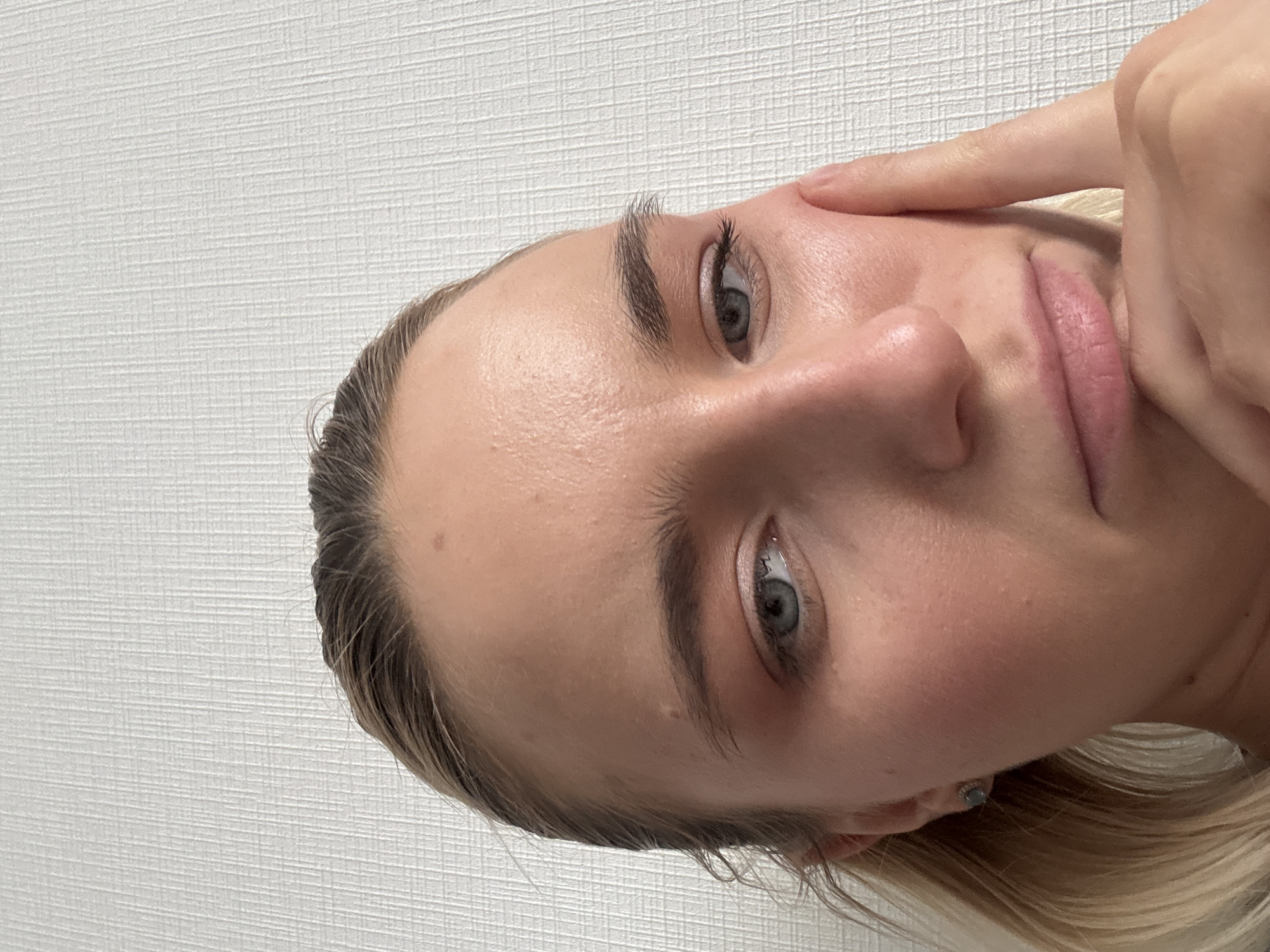 A close-up portrait of a young woman with light skin, light blonde hair, and grey eyes is presented against a white wall with a subtle texture. The woman's face is slightly turned towards the viewer's right. Her right index finger is touching the skin near her right temple. She has well-defined eyebrows and a subtle blush on her cheeks. The city and country are not evident in the scene.
