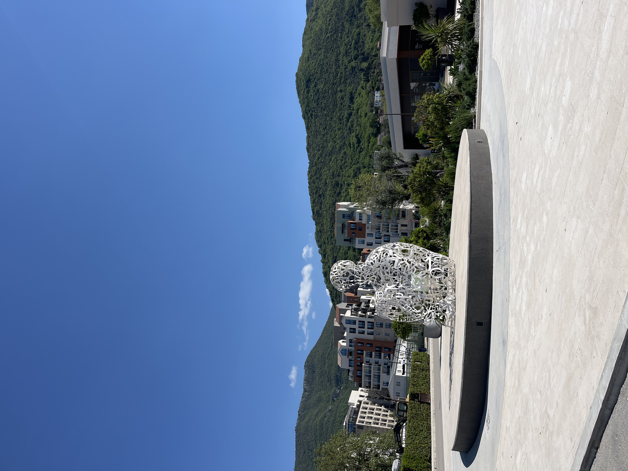 The image displays a vibrant outdoor scene in Tivat, Montenegro, under a clear blue sky. A prominent white sculpture, intricately formed from numerous interwoven letter-like shapes, occupies the foreground. The sculpture depicts a crouching or kneeling human-like figure, its openwork design creating a translucent effect. It is positioned on a wide, light-colored paved surface, likely a plaza or promenade. To the right of the sculpture, a dark, curved, low wall or edge, possibly part of a planter or fountain, casts a strong shadow.
In the mid-ground, a collection of modern buildings with various facades, including white, brown, and reddish-brown tones, are nestled at the foot of a large, verdant mountain range. The mountain's slopes are densely covered with green foliage, forming a lush backdrop. Scattered green bushes and trees add to the natural elements surrounding the architecture. The sky above is a bright, cloudless blue, save for a few small, wispy white clouds. The intense sunlight and sharp shadows indicate a midday or early afternoon setting. There is no discernible activity taking place, and no legible text other than the abstract shapes comprising the sculpture are visible. The scene combines contemporary art and architecture with stunning natural scenery.