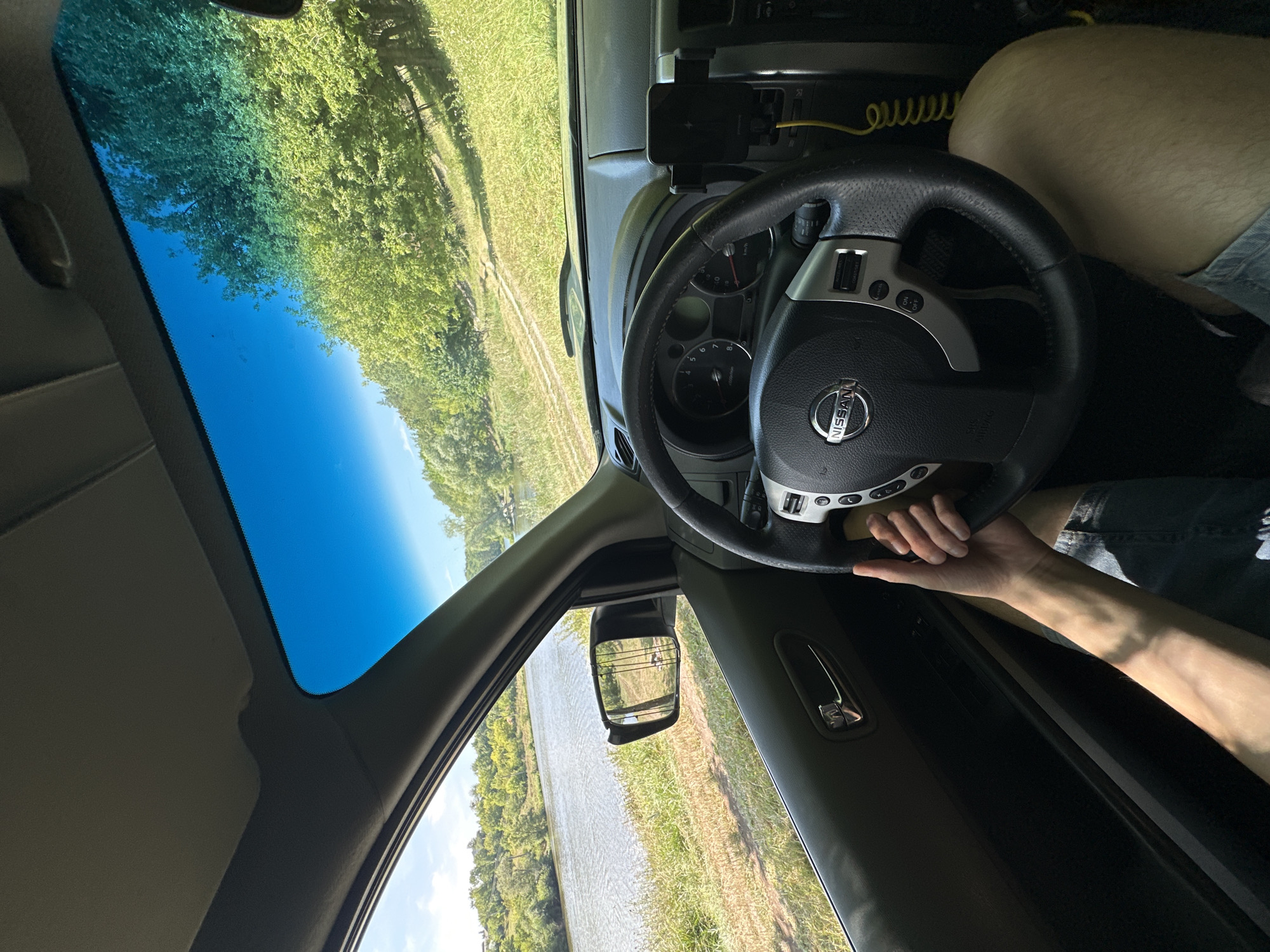 The image captures the interior of a Nissan X-Trail vehicle parked near a body of water, likely the Teteriv River, on the outskirts of Makiv, Ukraine. The driver's left hand is visible, resting on the steering wheel. The dashboard features the Nissan logo, and a phone mount is attached to the right side. Through the windshield and side window, a scenic view of the river and lush green foliage is observed. The sun shines brightly, casting a shadow of the sun visor on the windshield. The overall impression is one of a peaceful, outdoor setting.