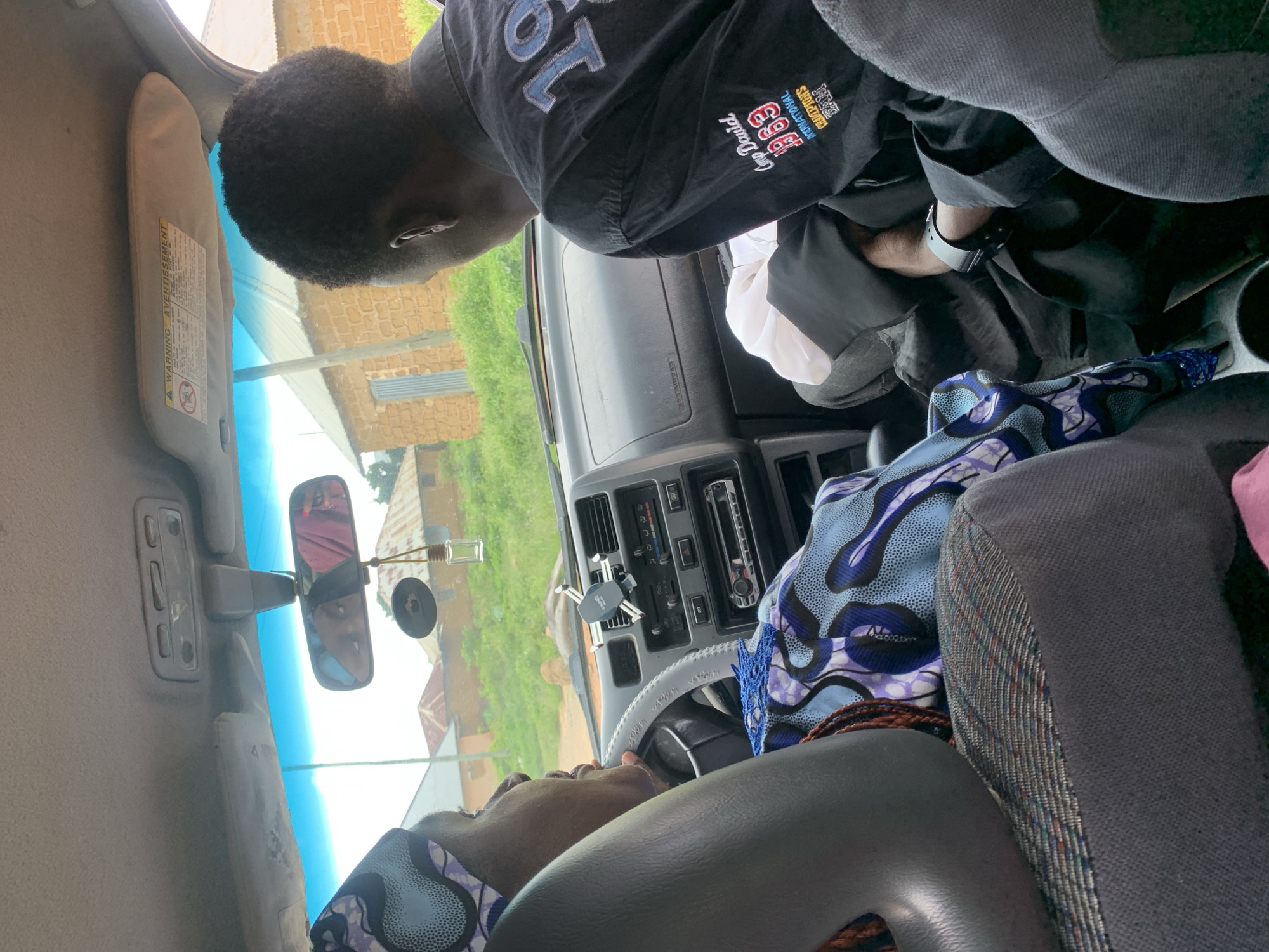 In an unknown city, two individuals are inside a car. The person in the driver's seat, a woman wearing a headscarf with a blue and purple pattern, is looking to her left. The passenger, a man in a black shirt with "Camp David 1969 International Champions Team" printed on the back, is looking forward. The car's interior includes a dashboard with a radio, air vents, and a mobile phone holder attached to the windshield. Outside the car, brick buildings and greenery are visible, suggesting a rural or suburban environment. The lighting indicates it is daytime, possibly overcast.