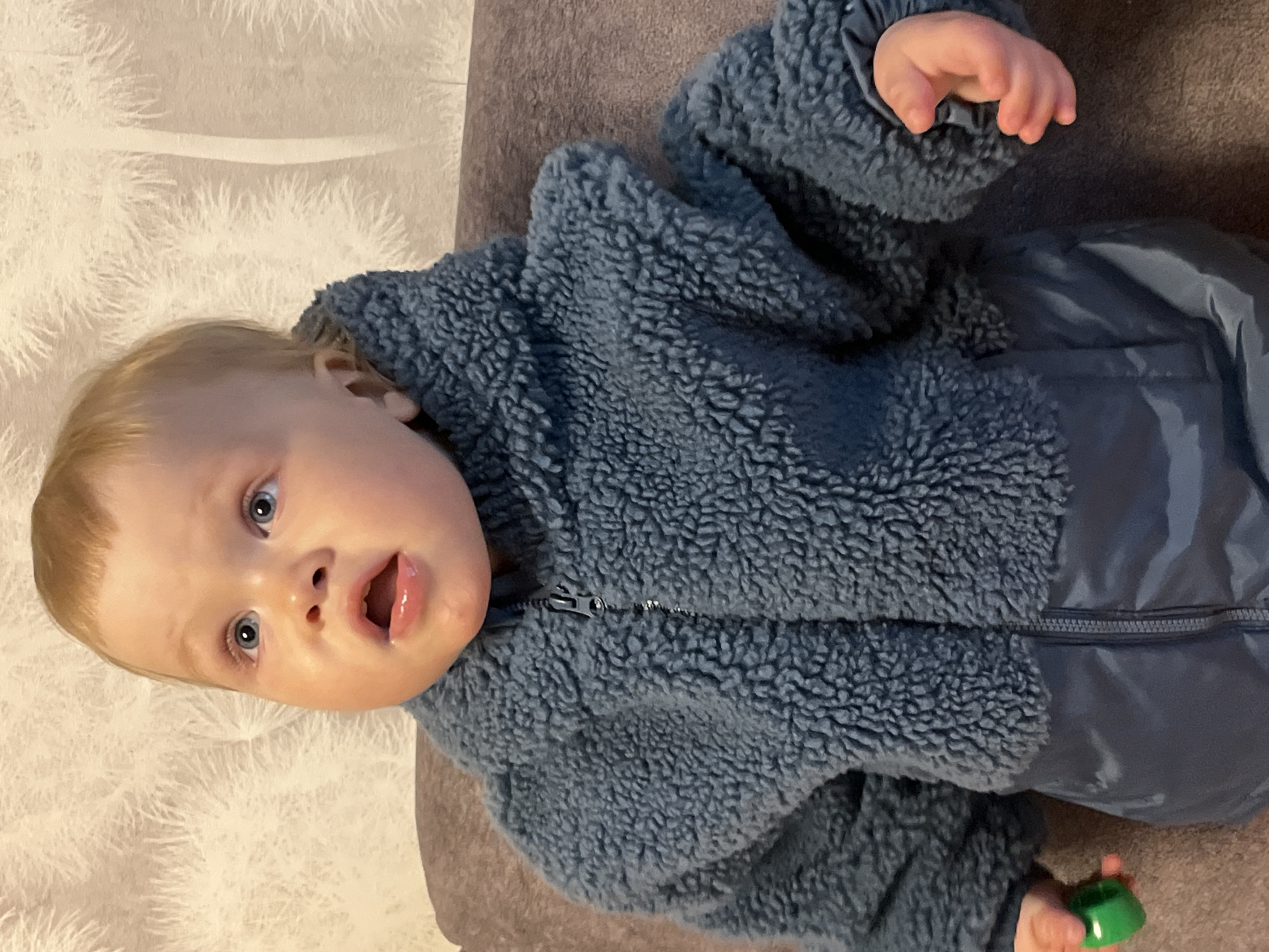 A baby with fair skin and blond hair is lying on a surface with a patterned white and light brown backdrop and a darker brown cushion or surface behind them. The baby is wearing a blue fuzzy jacket with a zipper and blue pants. The baby's mouth is open and they are looking upwards with blue eyes. A small green object is held in their left hand. It is impossible to confirm the location of the city of Chaikovsky, Russia with the provided image.