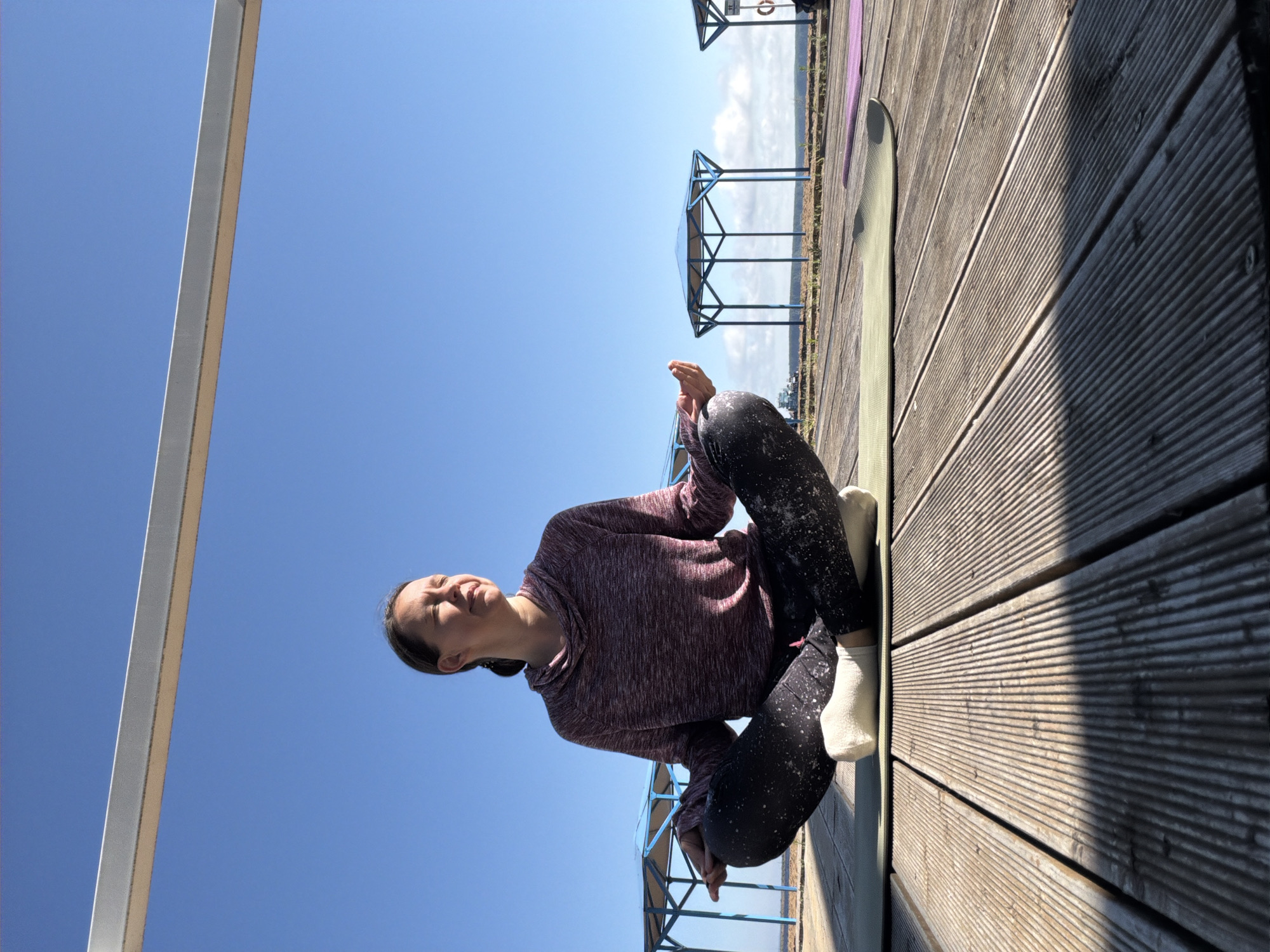 A woman sits in a lotus position on a yoga mat on a wooden deck. She is wearing a maroon hoodie and black leggings with white speckles, and white socks. Her eyes are closed and she appears to be meditating. In the background, there are blue gazebos and the sky is clear and blue. The wooden deck is made of parallel planks, and there are shadows cast by an overhead structure. The location appears to be an outdoor recreational area, possibly near a body of water. This scene is likely in Chaikovsky, Russia, given the context.