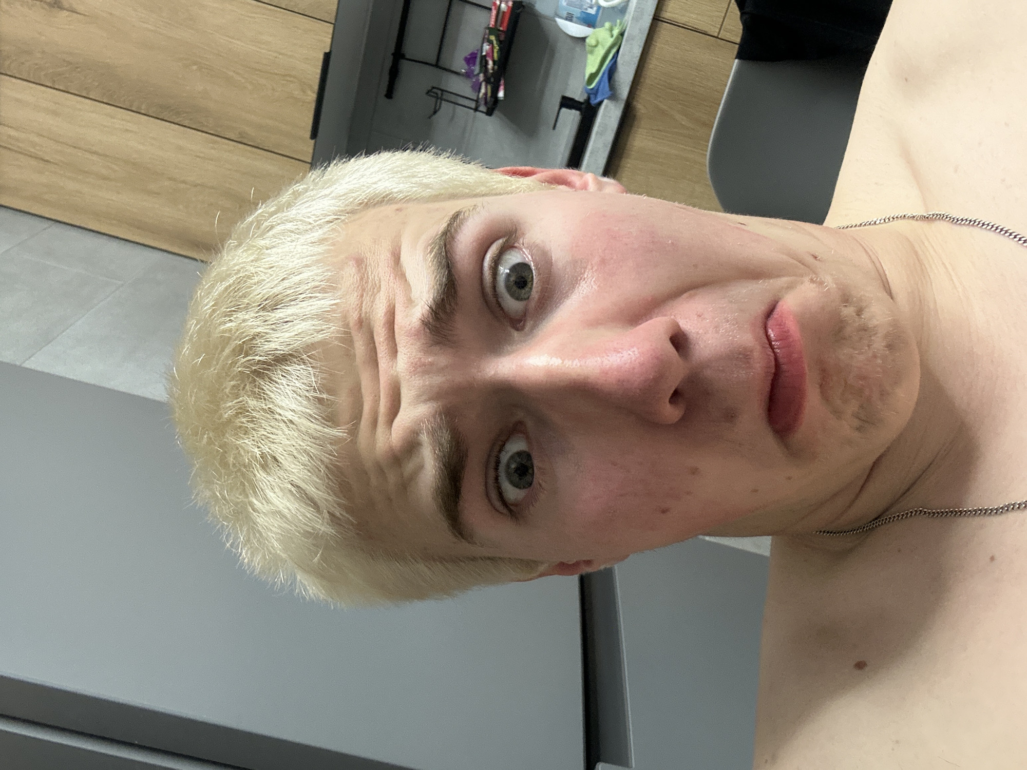 A close-up selfie captures a young man with short, bleached blonde hair and a shirtless torso, wearing a silver chain. He is looking directly at the camera with a highly exaggerated expression, characterized by wide, dilated eyes, a furrowed brow, and slightly parted lips, conveying surprise or humor. Light stubble is visible on his chin and jawline. The background elements suggest an indoor setting, possibly a bathroom or kitchen, featuring light brown wooden cabinetry or wall paneling, a gray tiled floor, and a mirrored or reflective surface in the upper background showing some indistinct household items on shelves. There is no visible text.