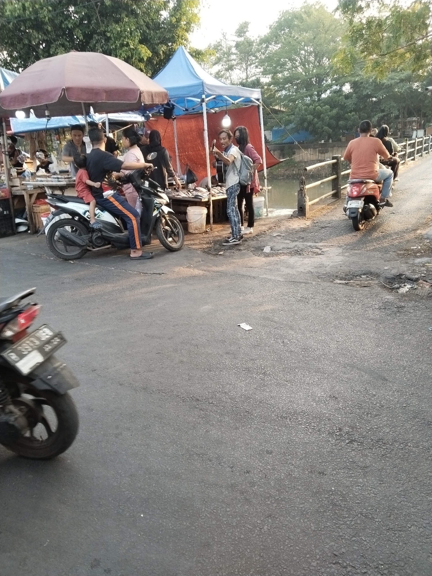 The image depicts a street scene at a market in West Jakarta, Indonesia. In the foreground, a motorcycle is parked, its license plate partially visible. The background features a bustling market with several people. A group of individuals are gathered around a stall, some interacting with merchandise, possibly food items. Two people are on a scooter, with a child sitting in front. Another motorcycle is seen with two riders further down the street, approaching a bridge. Tents and awnings of various colors, including maroon and blue, provide shade over market stalls. Trees and foliage are visible in the background, suggesting an outdoor setting. The ground is paved with asphalt, showing some signs of wear and tear. The lighting indicates it is late afternoon or early evening.