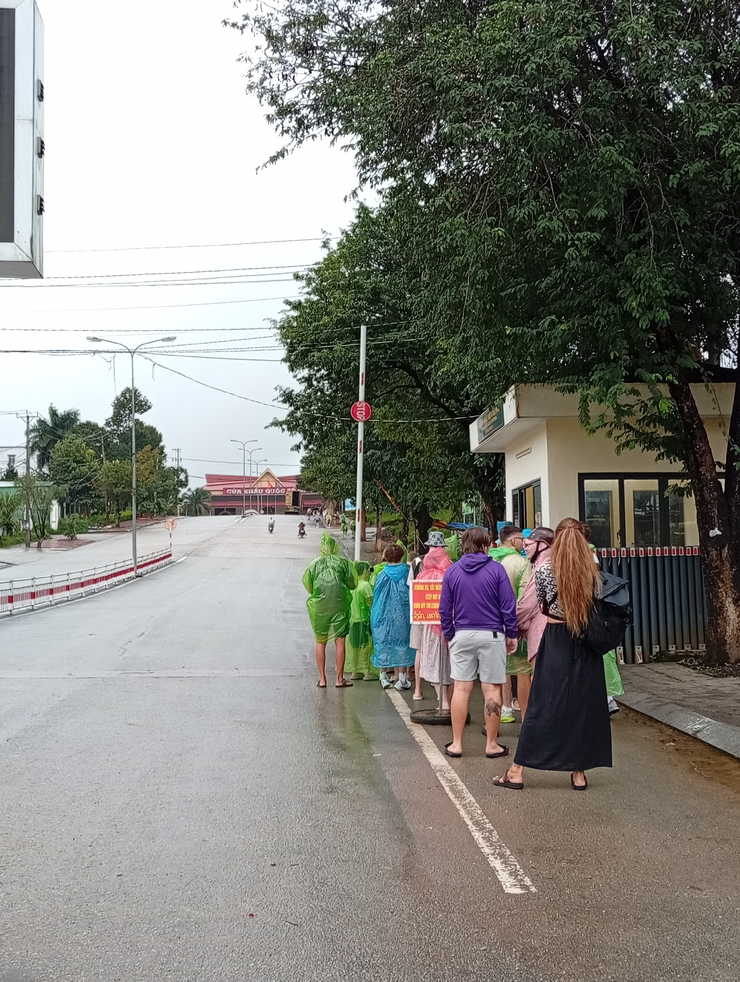 The image captures a group of people, some wearing bright raincoats, standing on a wet road in Bo Y, Vietnam, near a border crossing. A red and white barrier with a stop sign is visible to the right, indicating a checkpoint. In the background, a building with signage in Vietnamese and a red, peaked roof suggests a formal establishment, possibly related to border control or customs. Trees line the sides of the road, and the overcast sky indicates the weather is likely rainy or has recently rained. Several individuals are facing away from the camera, some holding a sign with text. Motorcycles are present on the road in the distance, suggesting vehicular traffic. The overall scene depicts a moment at a border area, with people possibly in transit or waiting for clearance.