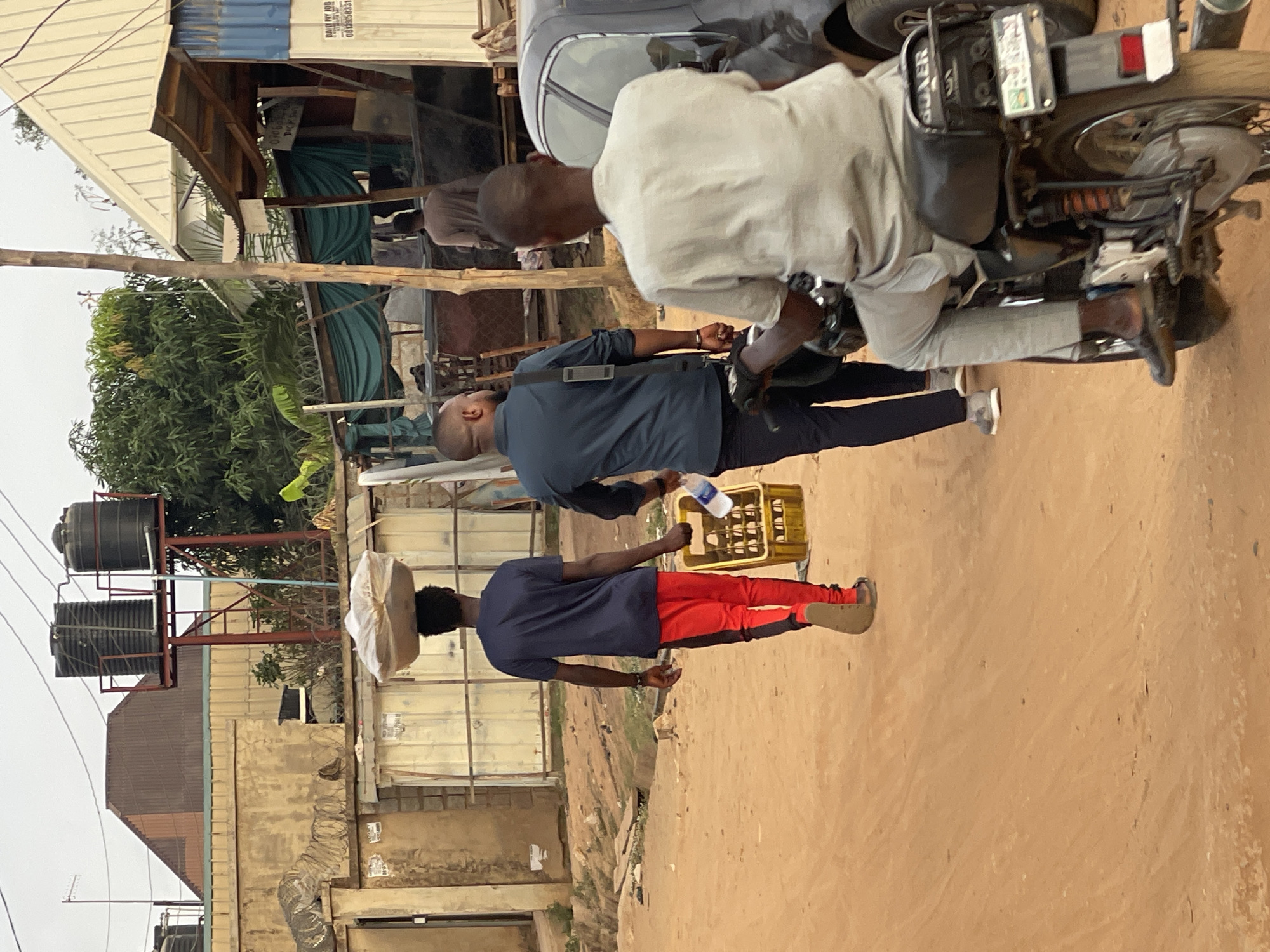 The scene depicts a street in Karmajiji, Nigeria. A man in a navy shirt and red pants is carrying a parcel on his head, walking away from the camera's perspective. Another man in a gray shirt and black pants follows behind him, holding a crate of drinks. A third man sits on a motorcycle parked next to them. The ground is dirt. Buildings and water tanks are visible in the background.