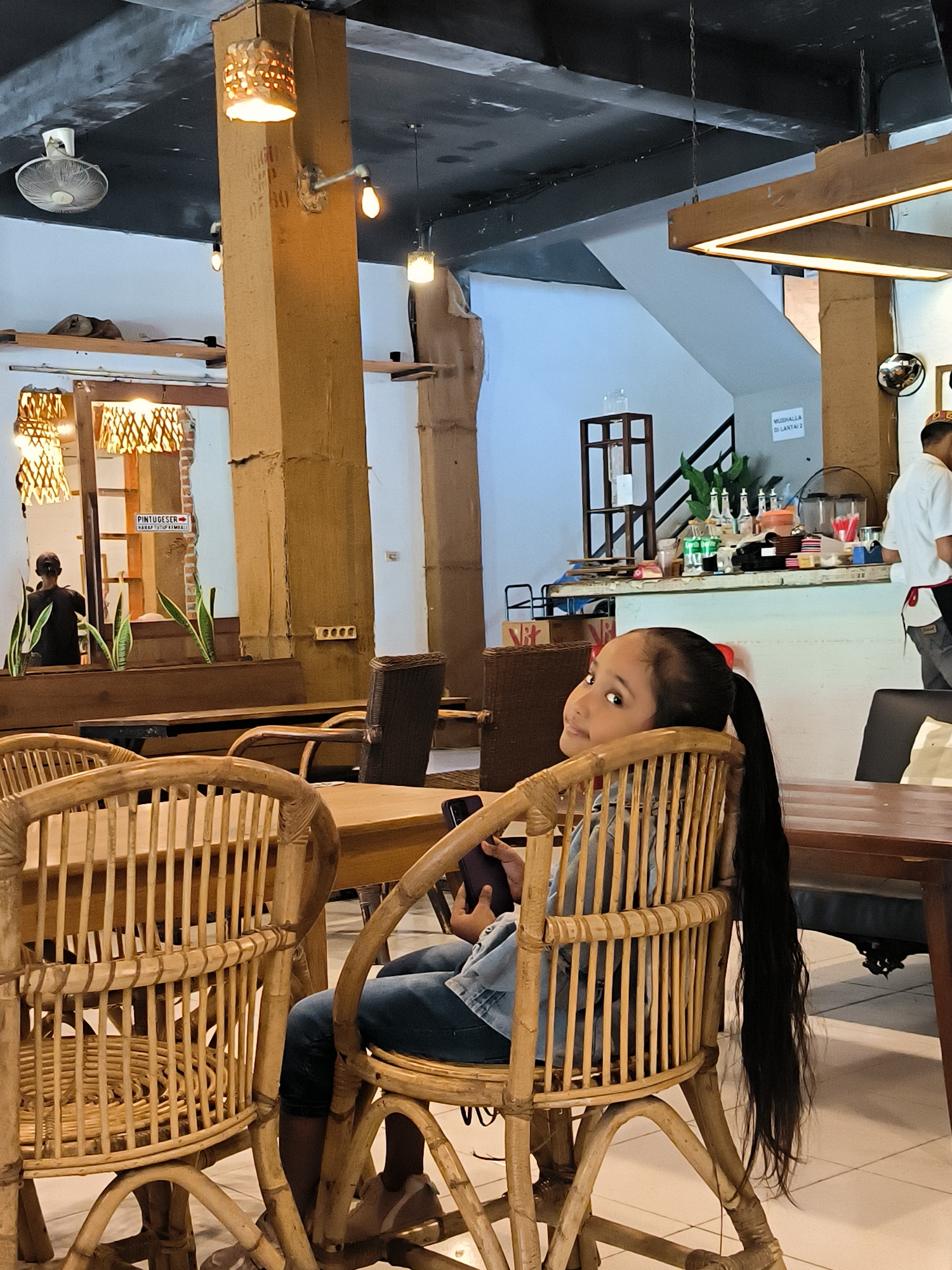 The image depicts the interior of a restaurant in Banda Aceh, Indonesia. In the foreground, a young girl with long black hair pulled back in a ponytail is seated in a rattan chair, looking back over her shoulder with a slight smile. She is wearing a denim jacket and jeans. The restaurant has a rustic ambiance with exposed beams, rattan furniture, and bamboo accents. Natural light filters in from windows and is supplemented by overhead pendant lights. In the background, a man in a white shirt and apron, possibly an employee, is visible near a counter. The restaurant appears to be a casual dining establishment, likely popular with locals and tourists.
