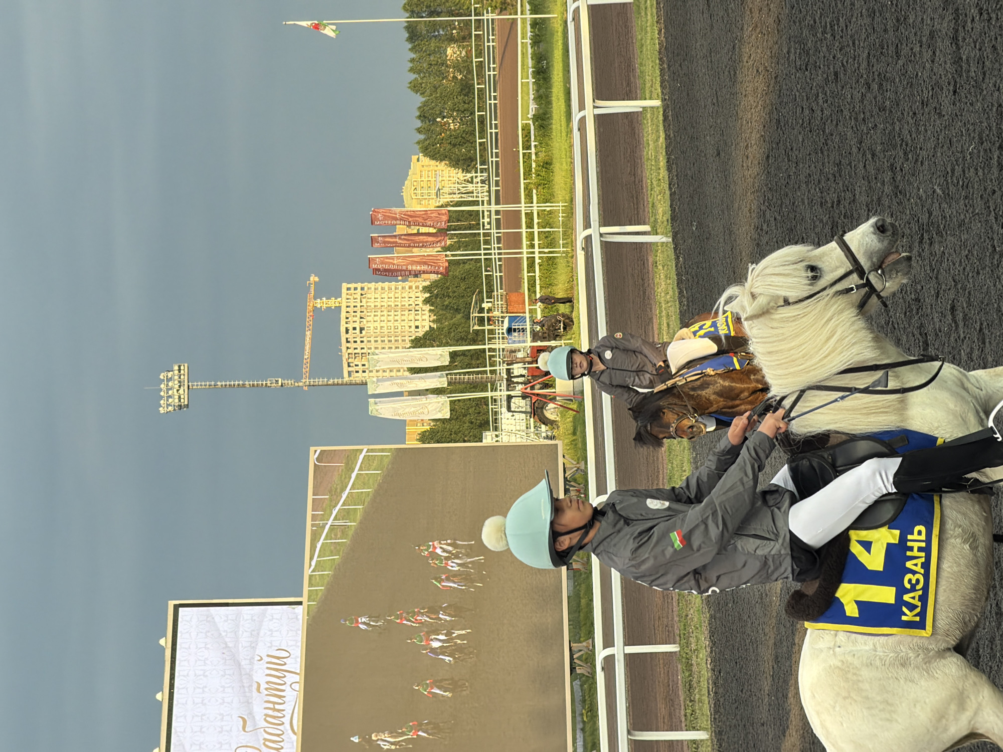 In a city, two children are riding ponies on a dirt track. The child in the foreground is riding a white pony and is wearing a light blue helmet, a gray jacket with a flag patch on the left sleeve, and white pants. The pony has a blue saddle blanket with the number "14" in yellow and the word "КАЗАНЬ" below it. The child in the background is riding a brown pony and is wearing a light blue helmet and a gray jacket. A large screen displays a horse race. In the background, there are flags, trees, buildings under construction, and a stadium light tower. The sky is overcast.