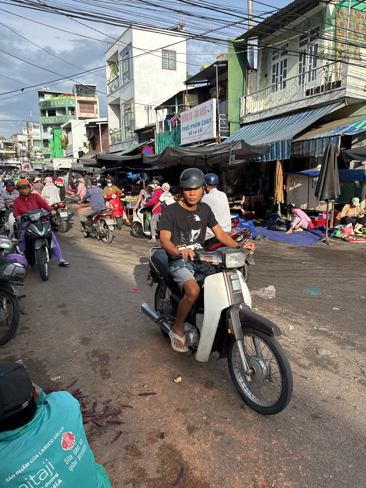 This is a street-level, eye-level, medium shot taken in Nha Trang, Vietnam, featuring a bustling market scene. The foreground is dominated by a man wearing a black helmet and a black t-shirt, riding a white and brown motorcycle. He is looking directly at the camera. Behind him, other motorcyclists and pedestrians navigate the crowded street. To the right, vendors are seen selling goods from stalls under awnings. The buildings in the background appear to be a mix of residential and commercial structures, typical of a Vietnamese city. The overall atmosphere suggests a lively and active marketplace.