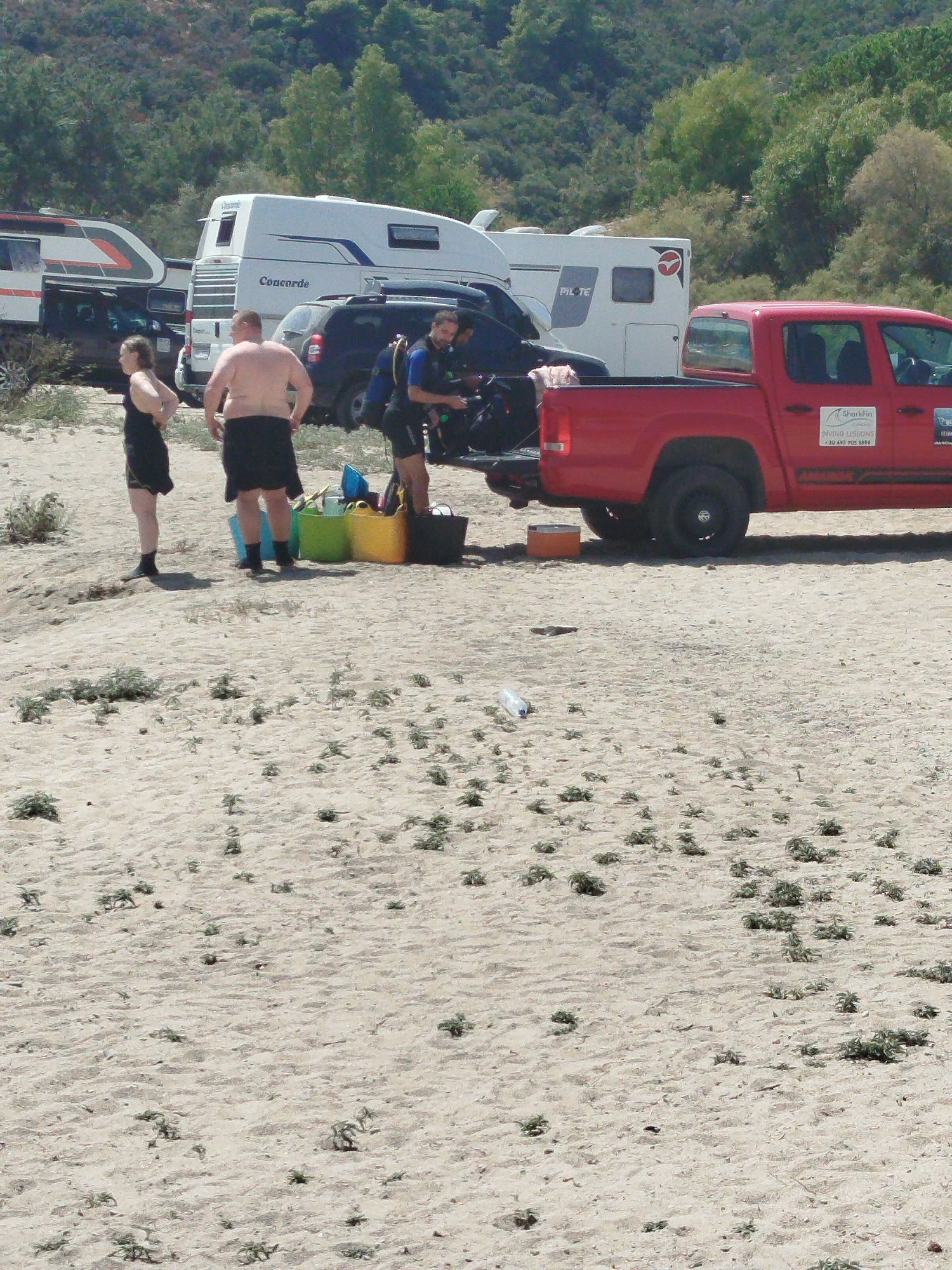 This image shows a sandy beach with sparse vegetation and several vehicles, including a red pickup truck, a white RV, and a black SUV. Three individuals are visible on the beach. One person, a woman in a black swimsuit, stands facing away from the camera. Another person, a man with no shirt and dark shorts, stands facing the camera. A third person, a man in diving gear, is partially obscured. Several large colorful buckets, a cooler, and diving equipment are present on the sand near the vehicles. The background consists of a dense, tree-covered hillside. The overall scene suggests an outdoor recreational activity, likely related to diving or water sports, taking place on a beach in Greece.