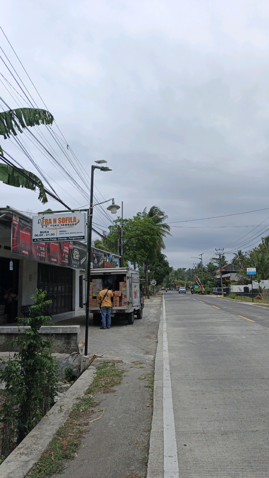 A street-level, medium shot captures a slightly overcast day in Cibenda, Indonesia, with a two-lane asphalt road running diagonally into the distance. On the left side of the road is a shop with a sign reading "FRA N SOFILA TOKO SEMBAKO" and "JL. RAYA TIMUR PARIGI, CIBENDA - PENGANDARAN". A person in a yellow shirt and blue jeans stands in front of a delivery truck, unloading boxes. The truck is open, revealing stacks of cardboard boxes. Power lines crisscross the sky above, and palm trees line the right side of the road. A small cat is visible on the concrete sidewalk in the foreground.
