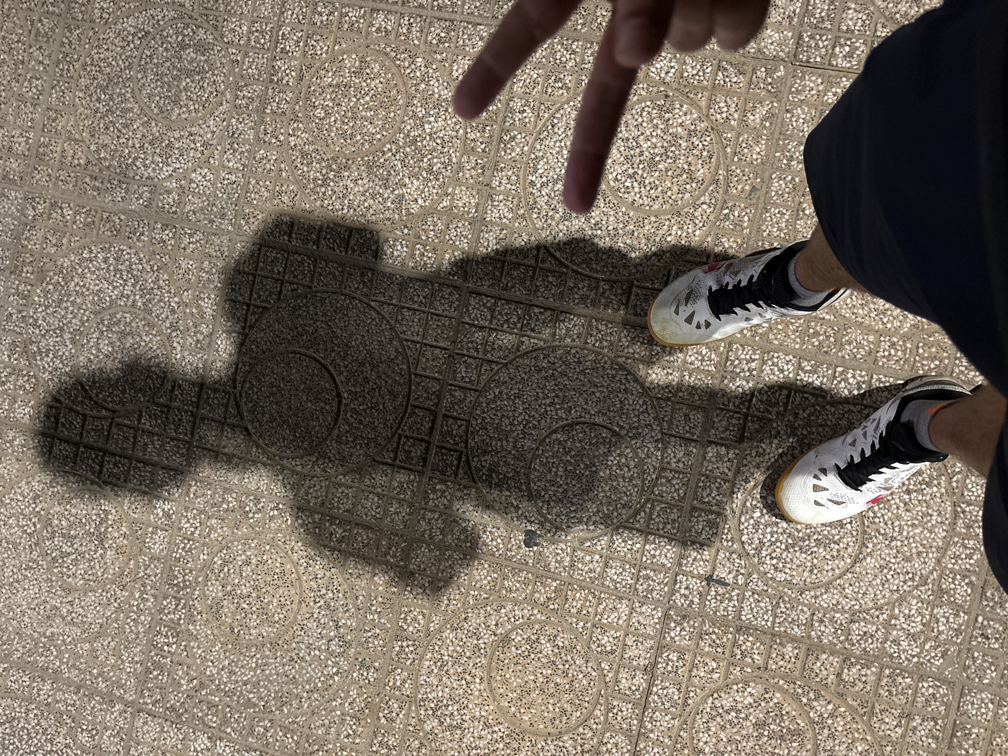The image shows a shadow of a person standing on a patterned pavement. The pavement is made of square tiles with circular patterns. The shadow is elongated and indistinct, suggesting the sun is at an angle. Two hands, one with fingers in a peace sign, are visible in the bottom right corner, belonging to the person casting the shadow. The person is wearing dark shorts and white athletic shoes. The scene appears to be outdoors on a sunny day.