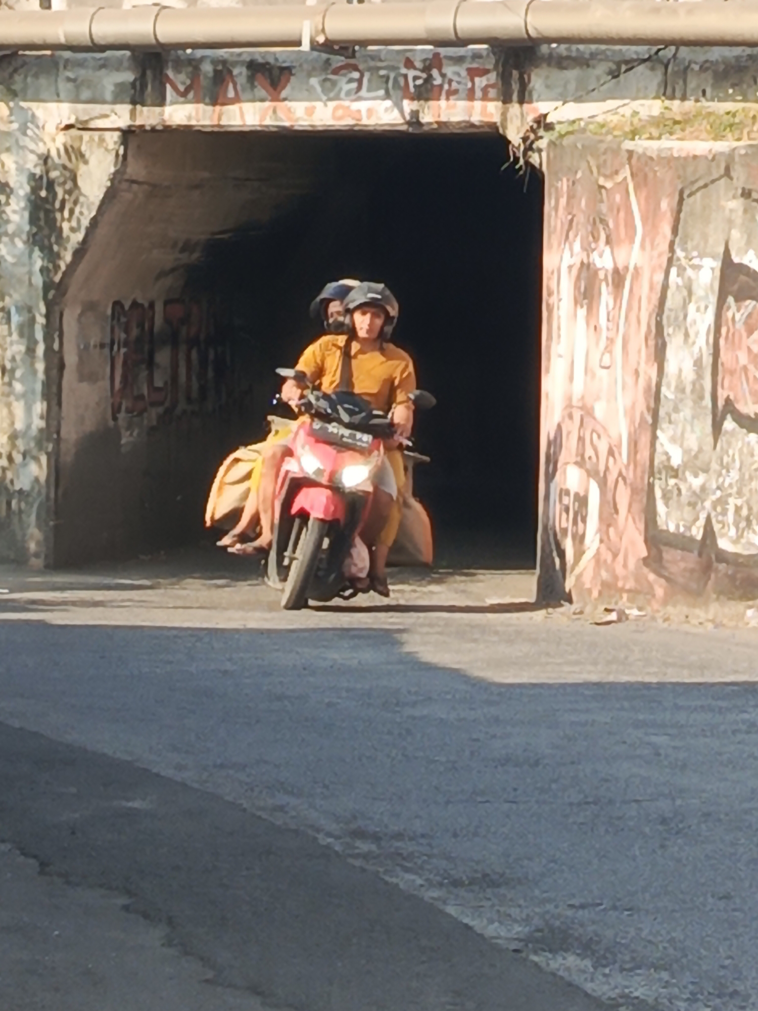 Two individuals are riding on a red motorcycle entering a dark tunnel. The driver, in the front, wears a yellow shirt and a dark helmet. The passenger, behind the driver, is partially obscured by the driver and also wears a helmet. The motorcycle's headlights are illuminated. The tunnel's entrance is framed by graffiti-covered concrete walls. The ground in front of the tunnel is paved, with a lighter colored road curving away to the right. The overall scene is lit by natural daylight.