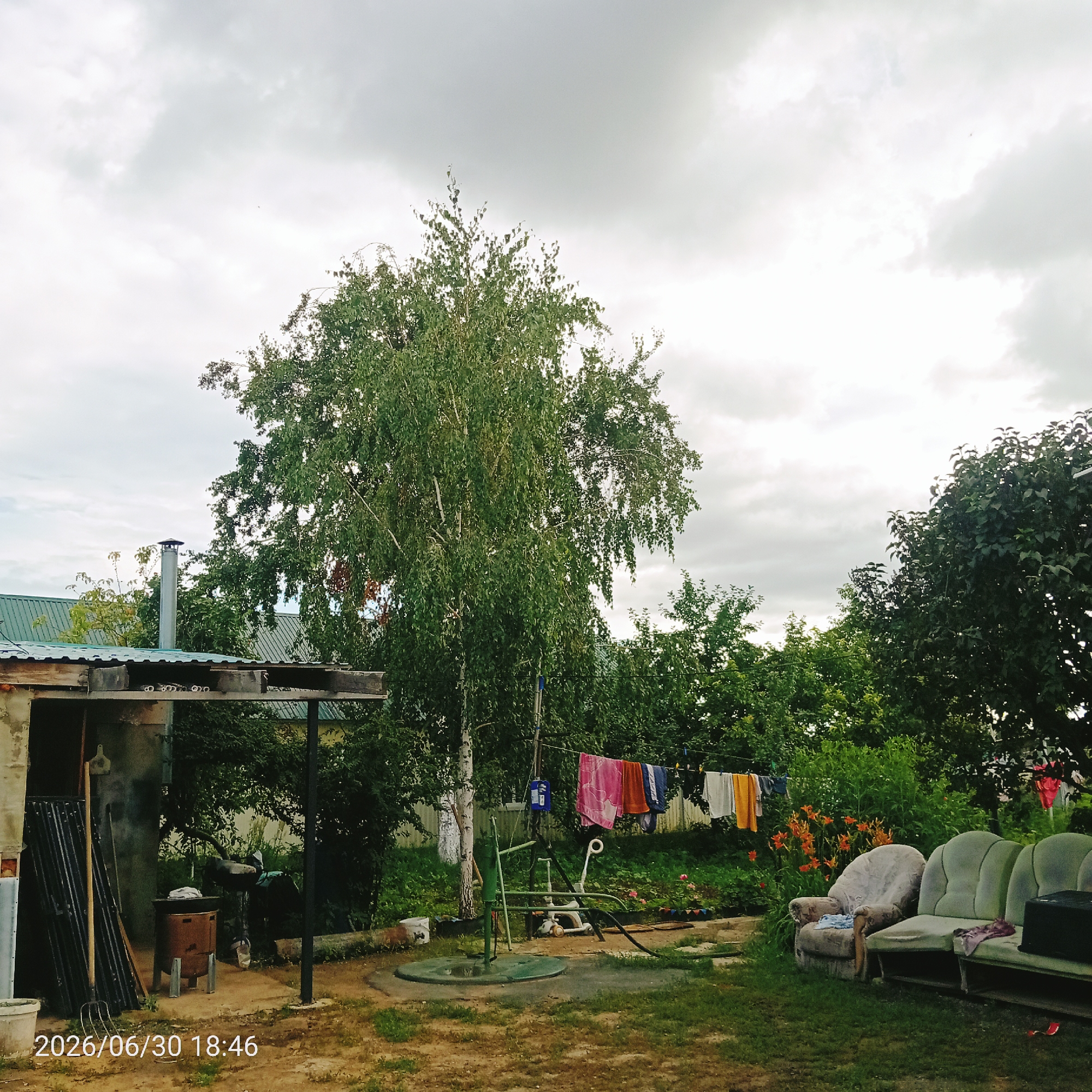 This media file depicts an outdoor scene, likely a backyard or garden area, on an overcast day. The prominent subjects include a tall birch tree in the center, a clothesline with various garments hanging to dry, a water well with a hand pump, and a small structure on the left that appears to be a shed or utility building. In the foreground, two outdoor chairs or sofas are visible, one of which has laundry draped over it.
The setting is rural or suburban, with dense greenery and trees forming a backdrop. The sky is filled with clouds, suggesting a cloudy or potentially rainy day. The presence of the clothesline and drying laundry implies a domestic activity. The water well and pump indicate a need for manual water retrieval. The overall atmosphere is one of a lived-in outdoor space.
Notable details include the clothes on the line, which are brightly colored, including pink, orange, and blue. The chairs/sofas appear weathered, adding to the rustic feel of the scene. The time of day is indicated by the visible text in the bottom left corner: "2026/06/30 18:46", suggesting it was taken late in the afternoon. The location is unknown, but the elements suggest a temperate climate. There are no people visible in the image. The text also includes a date and time stamp.