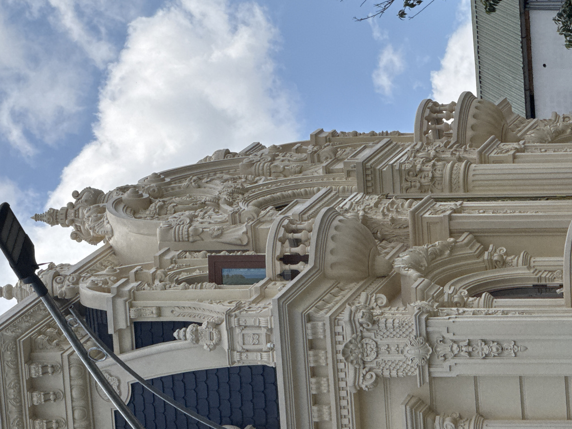 This is a close-up, eye-level, outdoor, medium shot of the elaborately ornamented facade of a cream-colored building, likely a French colonial-style villa, in Da Lat, Vietnam. The building features intricate stucco work, including carved floral motifs, column capitals, and decorative friezes. A dark blue, shingled roofline is visible on the left, meeting the ornate building. A small, open wooden-framed window is also visible on a balcony. The sky above is a bright blue with scattered white clouds. A street lamp extends from the top left corner of the frame, partially obscuring the building. A bare tree branch is visible in the upper right corner, and a green corrugated metal roof of another building is partially visible in the lower right. The architectural style suggests it is located in the historical district of Da Lat, known for its French colonial architecture. There are no people visible in the image.