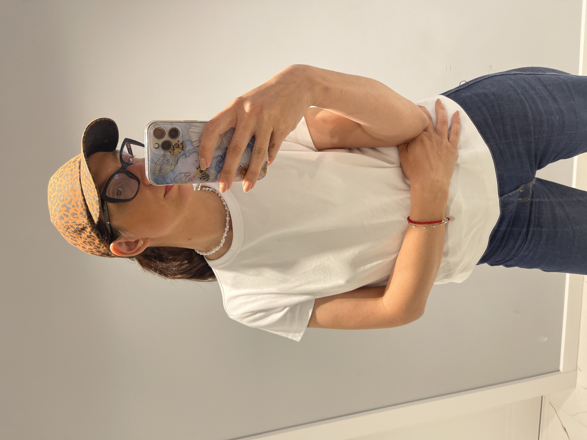 A woman with short brown hair is taking a selfie in front of a white wall. She is wearing a white t-shirt, blue jeans, a pearl necklace, a red bracelet, and black-framed glasses. She is also wearing a leopard print baseball cap. Her phone has a clear case with a bee design on the back. The location of the photo is unknown, and there is no indication it was taken in Sofia, Bulgaria.
