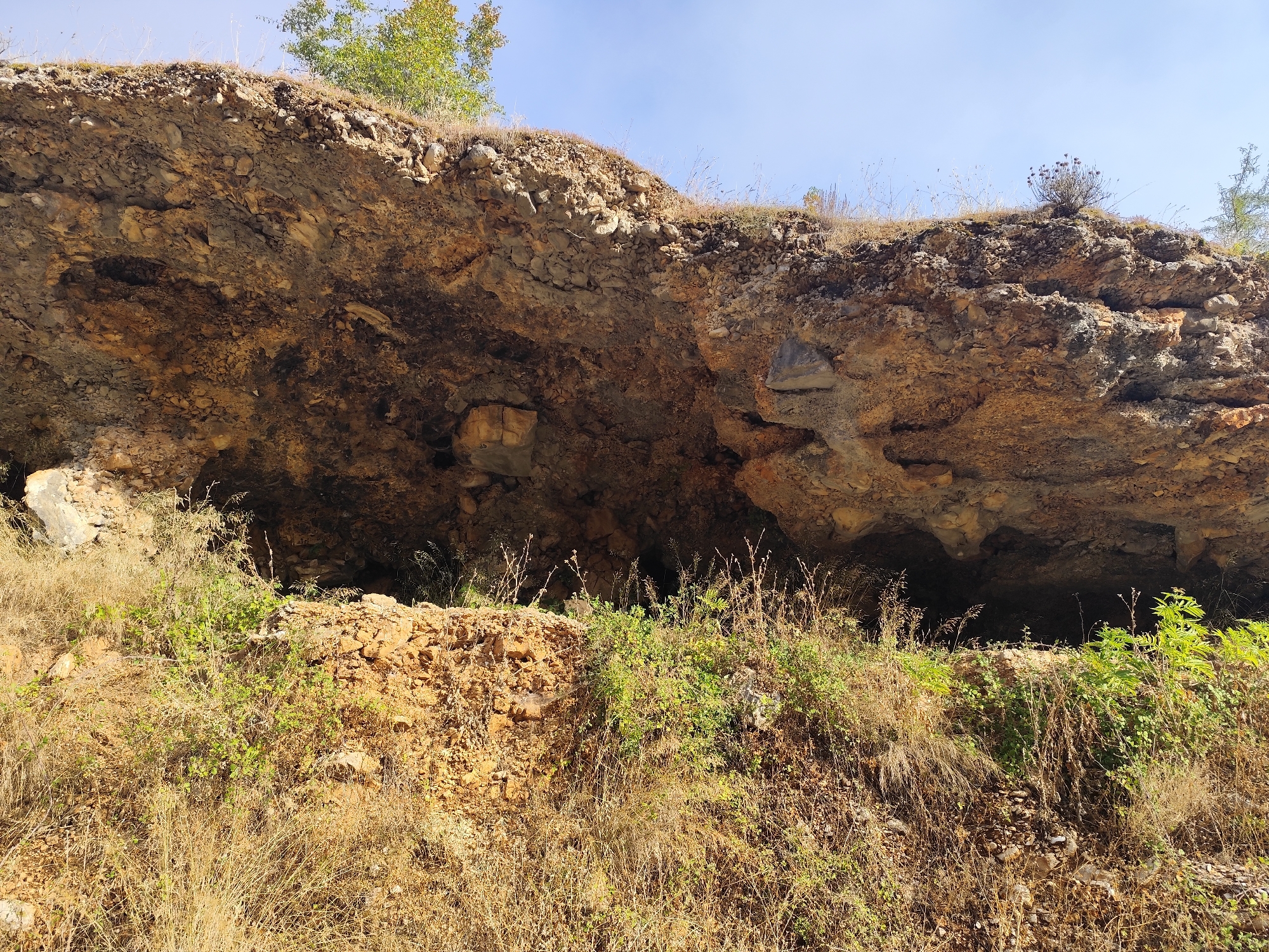 The image depicts a natural rock overhang or small cave on a hillside. The rock formation is predominantly earthy brown and grey with visible layers and mineral deposits. The top of the overhang is covered with dry grass, sparse vegetation, and some rocks. The entrance to the overhang is dark and appears to lead into a deeper recess. The foreground consists of a slope covered in dry, golden grass and some green bushes and saplings. The sky above is a clear blue. The scene is located in Dropull i Sipërm, Albania, a region known for its natural landscapes. There are no people or animals visible in the image. The overall environment suggests a dry, rural or natural setting.