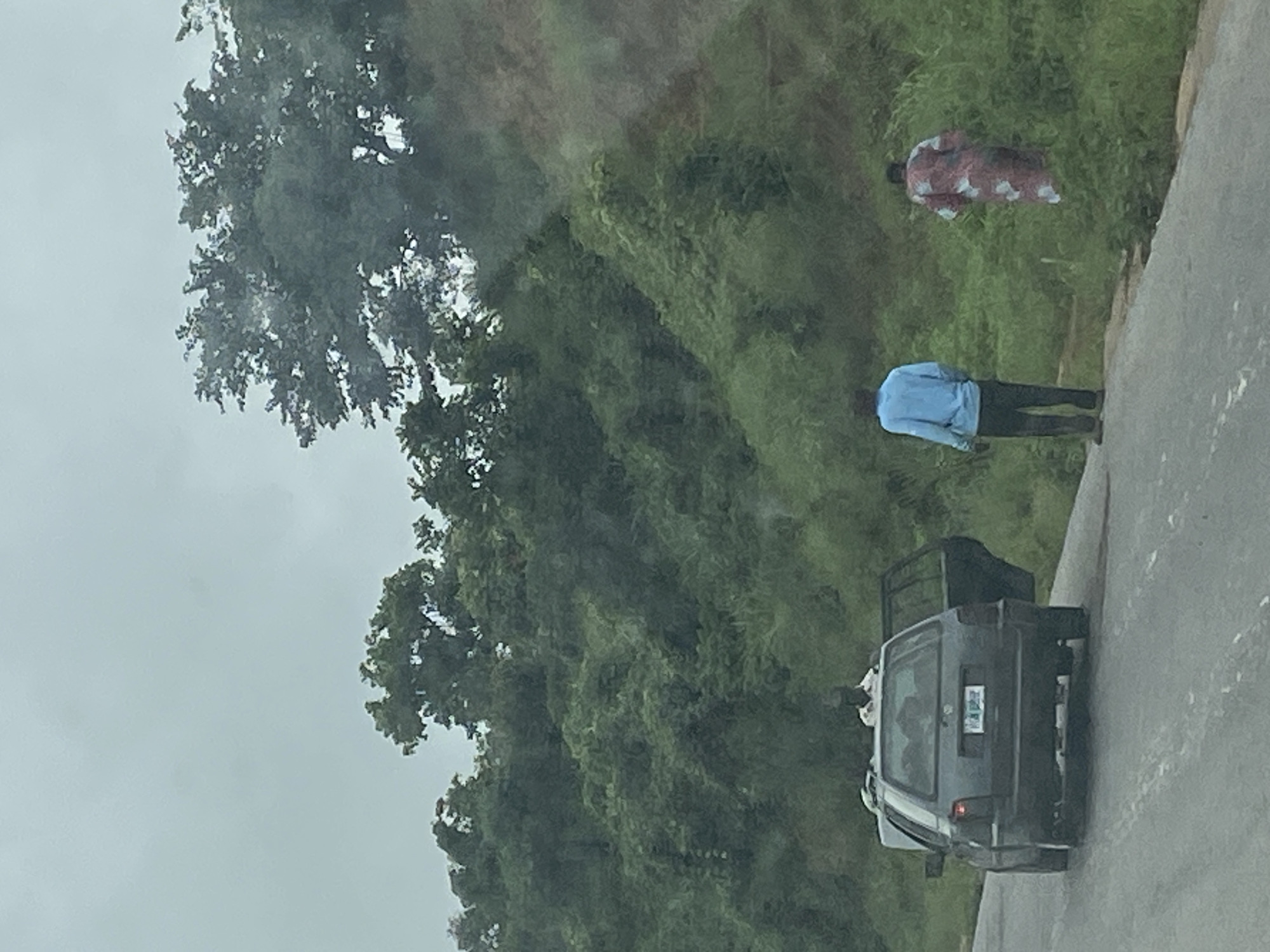 In Assob Hausa, Nigeria, a dark gray station wagon is parked on the side of a paved road with its rear hatch and driver's side door open. A person wearing a white shirt and dark headwear is visible from the waist up, standing inside the open rear of the vehicle. Further down the road, a person dressed in a blue long-sleeved shirt and dark pants stands facing away from the camera, with their back to the car. To the right of this person, a second individual is seen from behind, wearing a long, floral patterned robe, standing in tall green grass. The background consists of a dense, green, overgrown hillside with numerous trees and foliage under a cloudy sky.