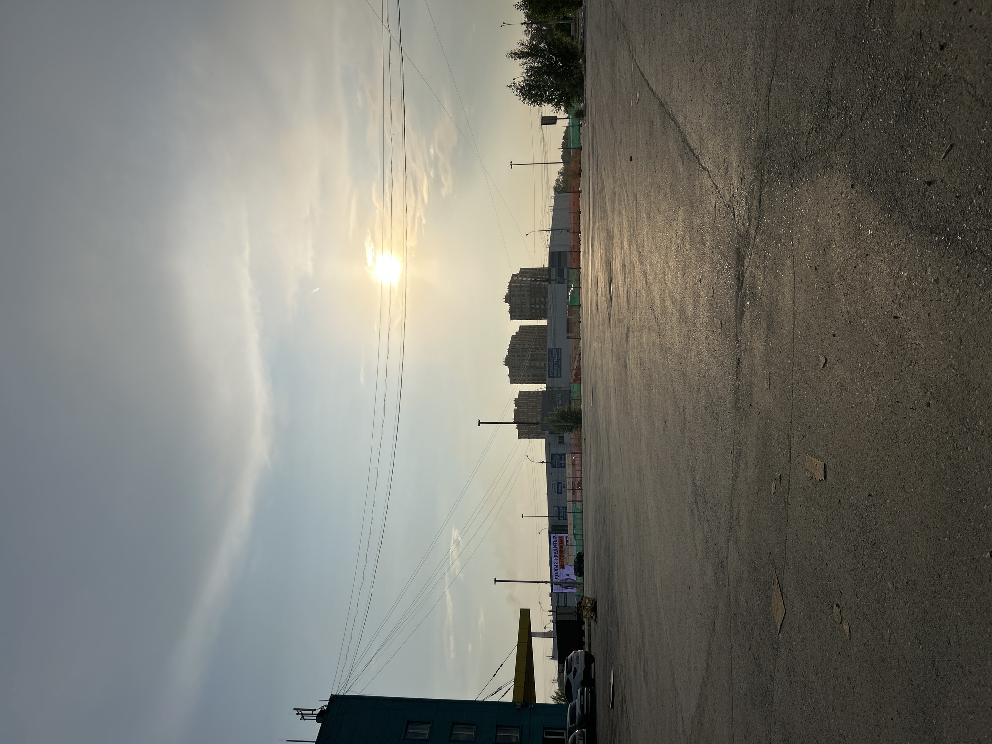The image shows an exterior shot of an urban environment during the day. The sun is low in the sky, suggesting it is either early morning or late afternoon, casting a warm, bright light. The sky is mostly clear with some wispy clouds.
In the foreground, there is a paved asphalt surface, possibly a road or parking lot, with some cracks and debris visible. To the left, part of a teal-colored building with windows can be seen. In the mid-ground, several tall apartment buildings are visible, suggesting a residential or mixed-use area. There are also power lines stretching across the sky. Further back, more buildings and trees are present, with some construction fencing and structures indicating ongoing development or renovation.
A large billboard with text is visible, featuring a prominent advertisement. The text on the billboard, though partially obscured and small, appears to be in Cyrillic script. It reads: "ДОЛГИ? КРЕДИТЫ? МЫ ПОМОЖЕМ!" which translates to "DEBTS? CREDITS? WE WILL HELP!".
The overall impression is of a typical city scene, possibly in Eastern Europe or a region using Cyrillic, with a mix of older and newer constructions. The lighting suggests a pleasant day, with the sun highlighting the textures of the asphalt and the silhouettes of the buildings. There are no people visibly depicted in the image.