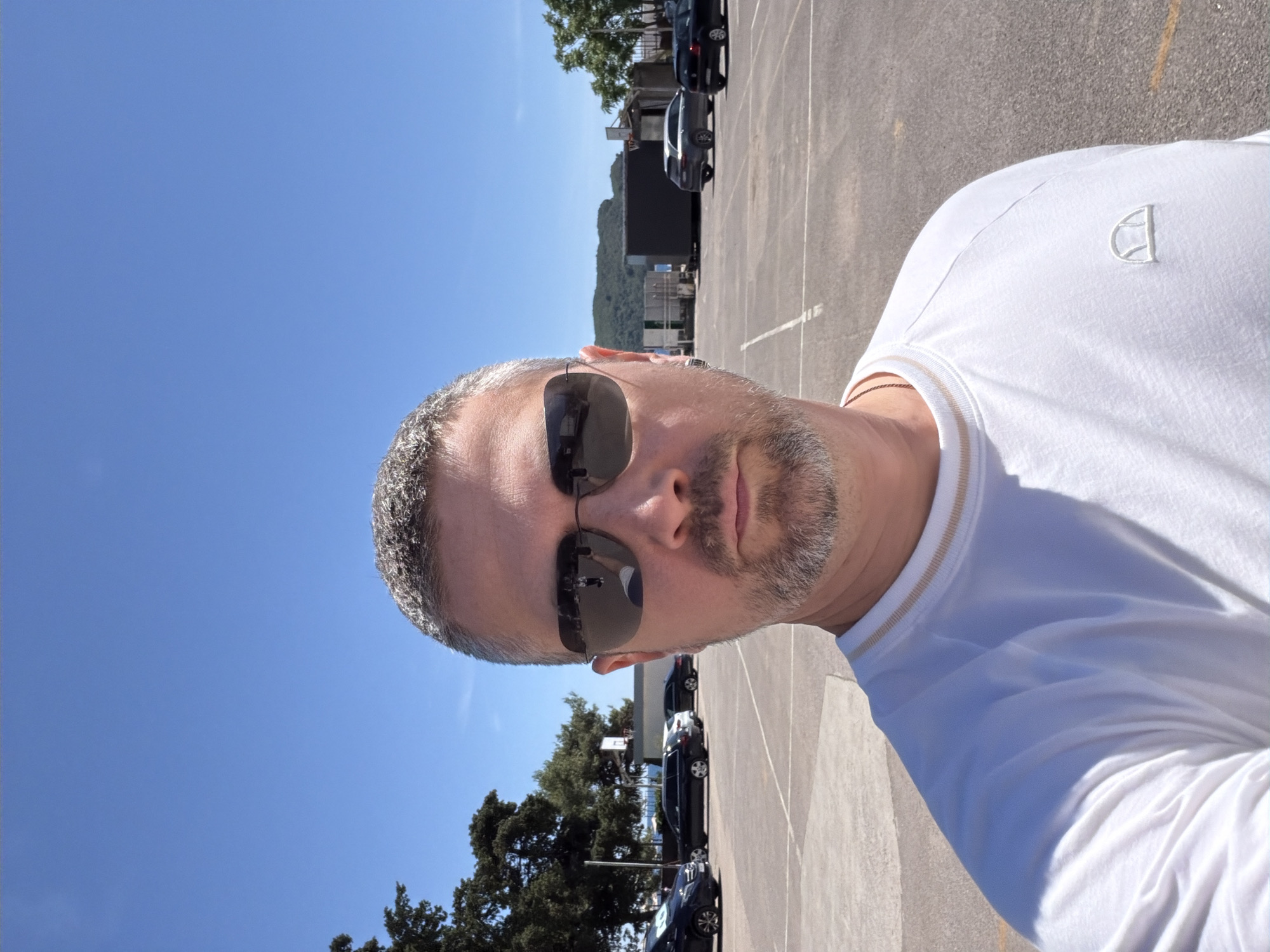 A selfie, shot outdoors under a clear blue sky. The subject, a Caucasian male with gray hair and a short beard, wears sunglasses and a white t-shirt with a logo. He appears to be standing in a parking lot with cars parked in the background. The location seems to be in Budva, Montenegro, based on the landscape and architecture.