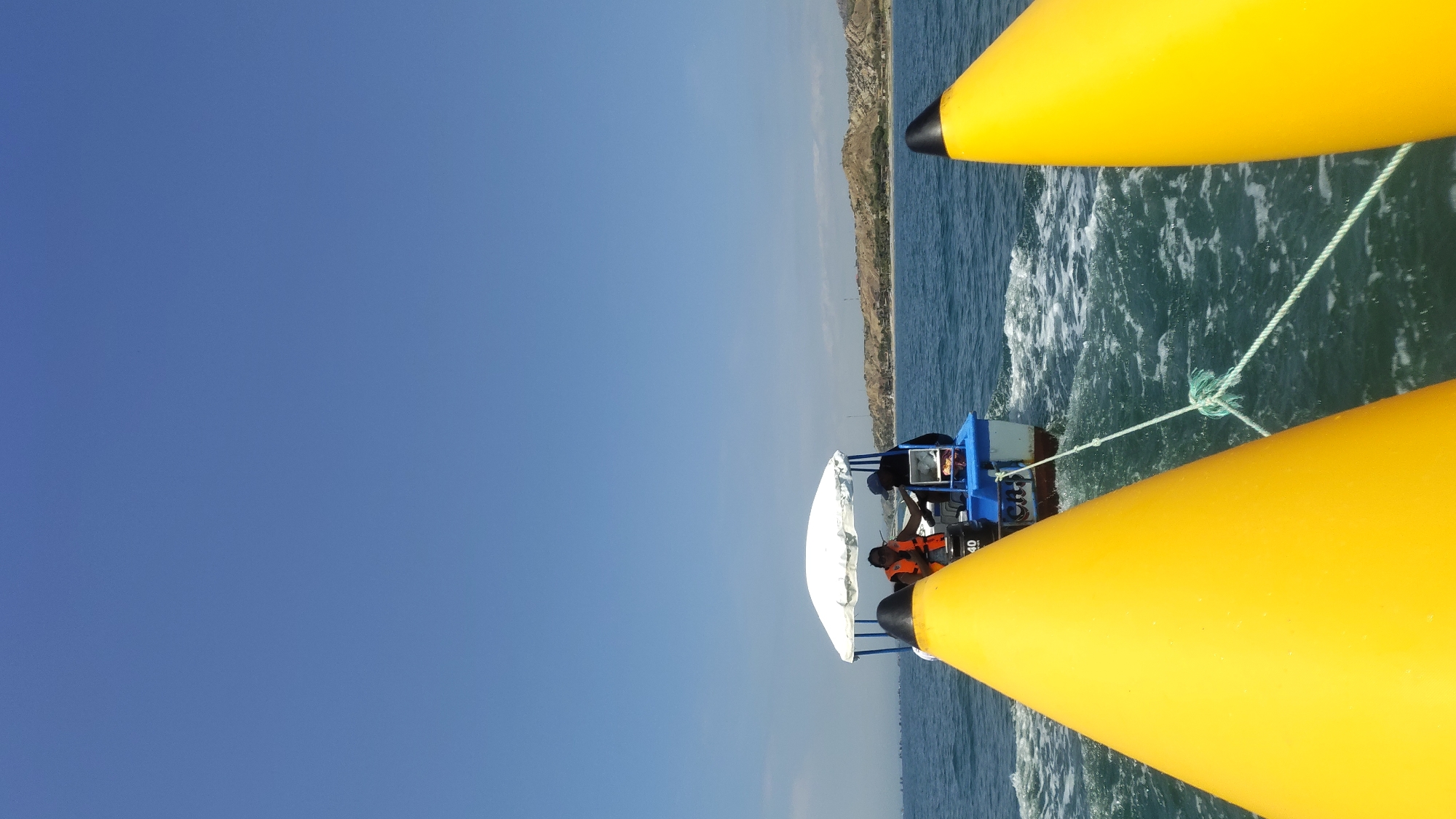 A boat with two passengers wearing life vests is being pulled behind a catamaran with yellow pontoons. A white and silver covering sits above the passenger area of the boat. The catamaran is moving through choppy, dark blue water, and the wake is visible between the pontoons. A light green rope is connecting the boat to the catamaran. In the background, there is a strip of land with buildings. The sky is a clear, bright blue. The location is Nuevo Cancas, Peru.