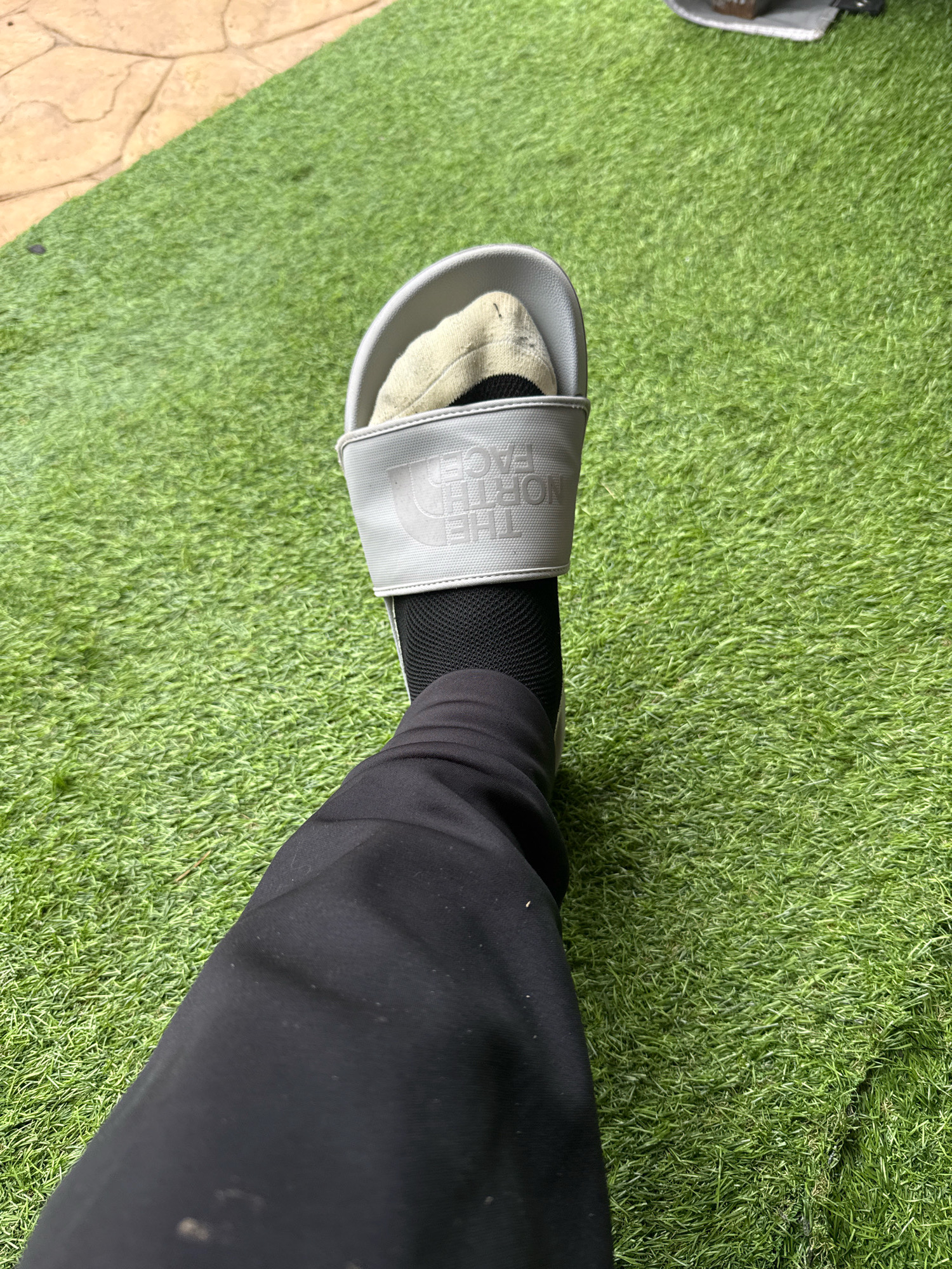 A first-person perspective shot focuses on a left foot clad in a white North Face slide with a beige sock peeking out and a black sock underneath, resting on artificial grass. The foot is also covered by a black pant leg. The background features a section of stone paving. No specific location in Colchester, United Kingdom can be identified.