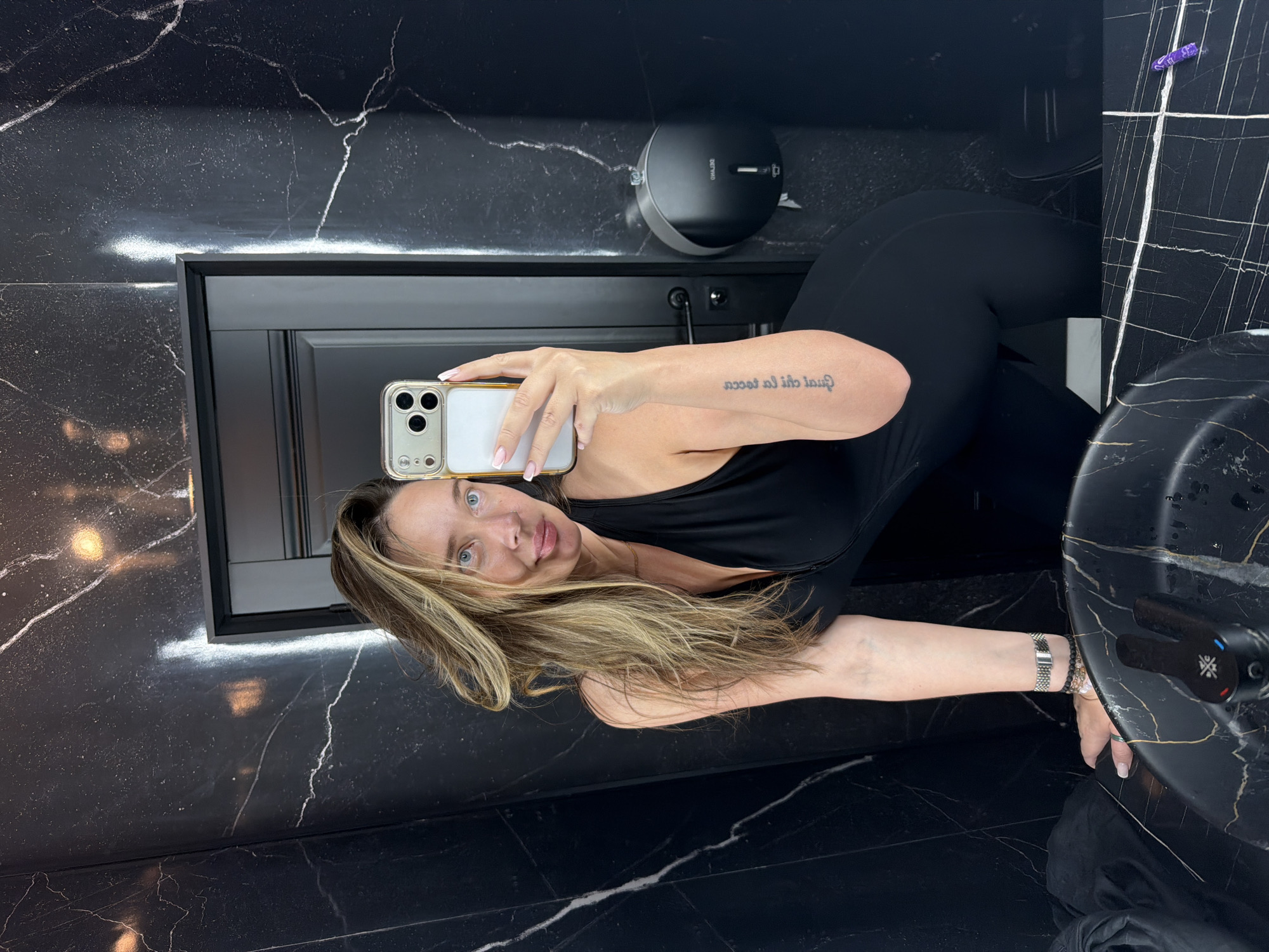 The media file shows a woman taking a mirror selfie in a bathroom with black marble walls and counters. She is wearing a black athletic outfit. The woman has long blonde hair and is looking at the camera. She has a tattoo on her arm that reads "Quoi au la tecca". She is also wearing jewelry on her wrist. The bathroom appears to be modern and luxurious. The time of day is not specified, but the lighting suggests it is indoors. The location is Moscow, Russia.