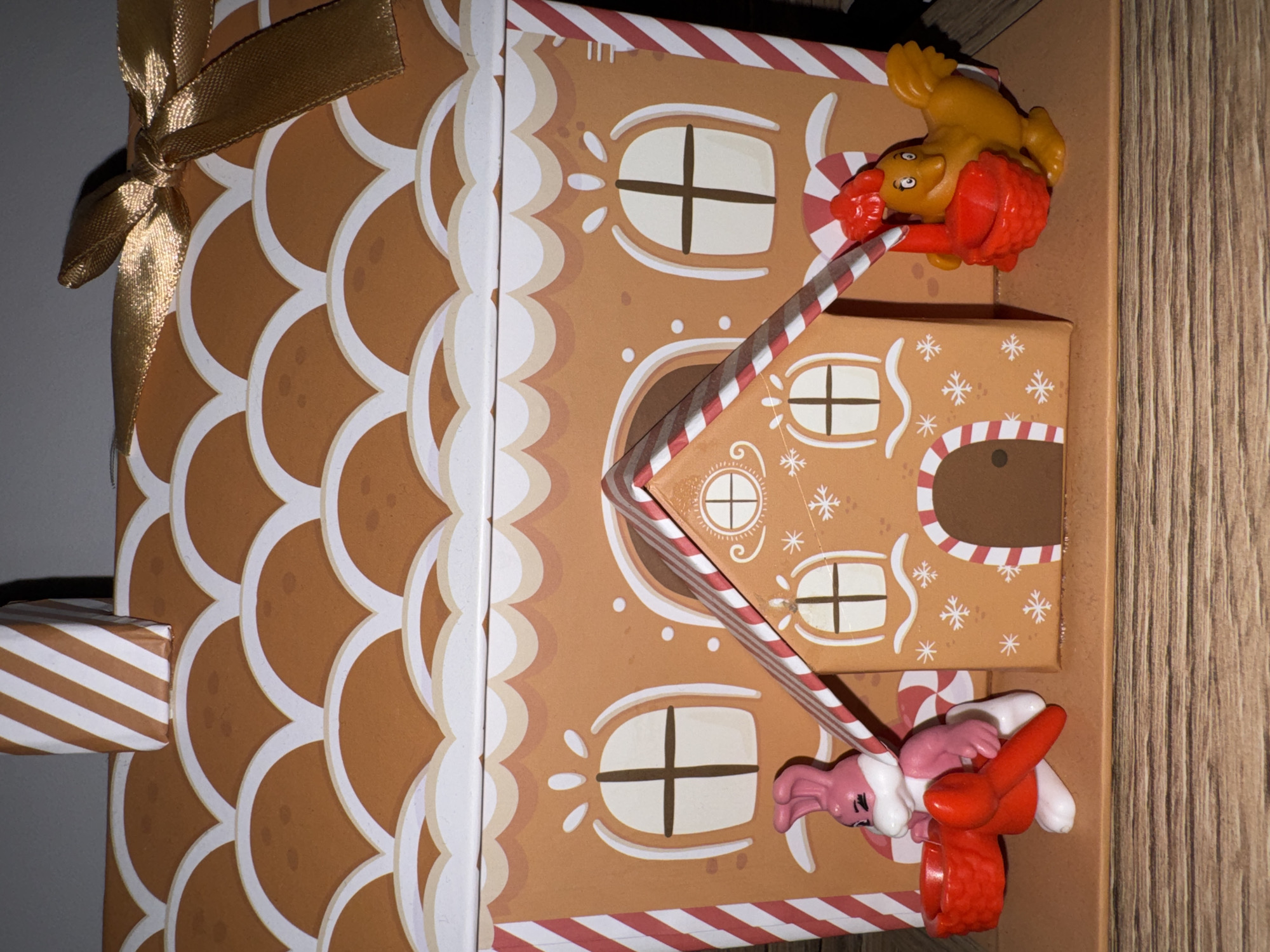 This image captures a festive, gingerbread house-themed item, likely an advent calendar or a gift box, resting on its side. The main subject is a brown cardboard structure decorated to resemble a gingerbread house, complete with white scalloped "icing" borders, various window designs with cross panes, and scattered white "snowflakes" and dots. Red and white striped candy cane patterns adorn the edges of the "roof" and a diagonal element across the facade.
A golden satin ribbon tied into a bow is visible on the left side of the item, suggesting it's either a decorative element or part of its closure mechanism. On the right, a wooden surface serves as the background.
Two small plastic toy figures are attached to the gingerbread house. The upper figure is predominantly yellow with red elements, resembling a bird-like character, possibly holding a red object like a watering can or scoop. It is positioned as if sliding down or hanging from one of the candy cane patterned structures. The lower figure is pink and white, appearing to be a rabbit or bunny, also holding onto a candy cane element. Both figures are stylized and cartoonish.
A small, brown and white striped "gift" box element protrudes from the left side of the main gingerbread house structure. No discernible text or branding is visible on the item or the figures. The scene evokes a playful and holiday-themed atmosphere.