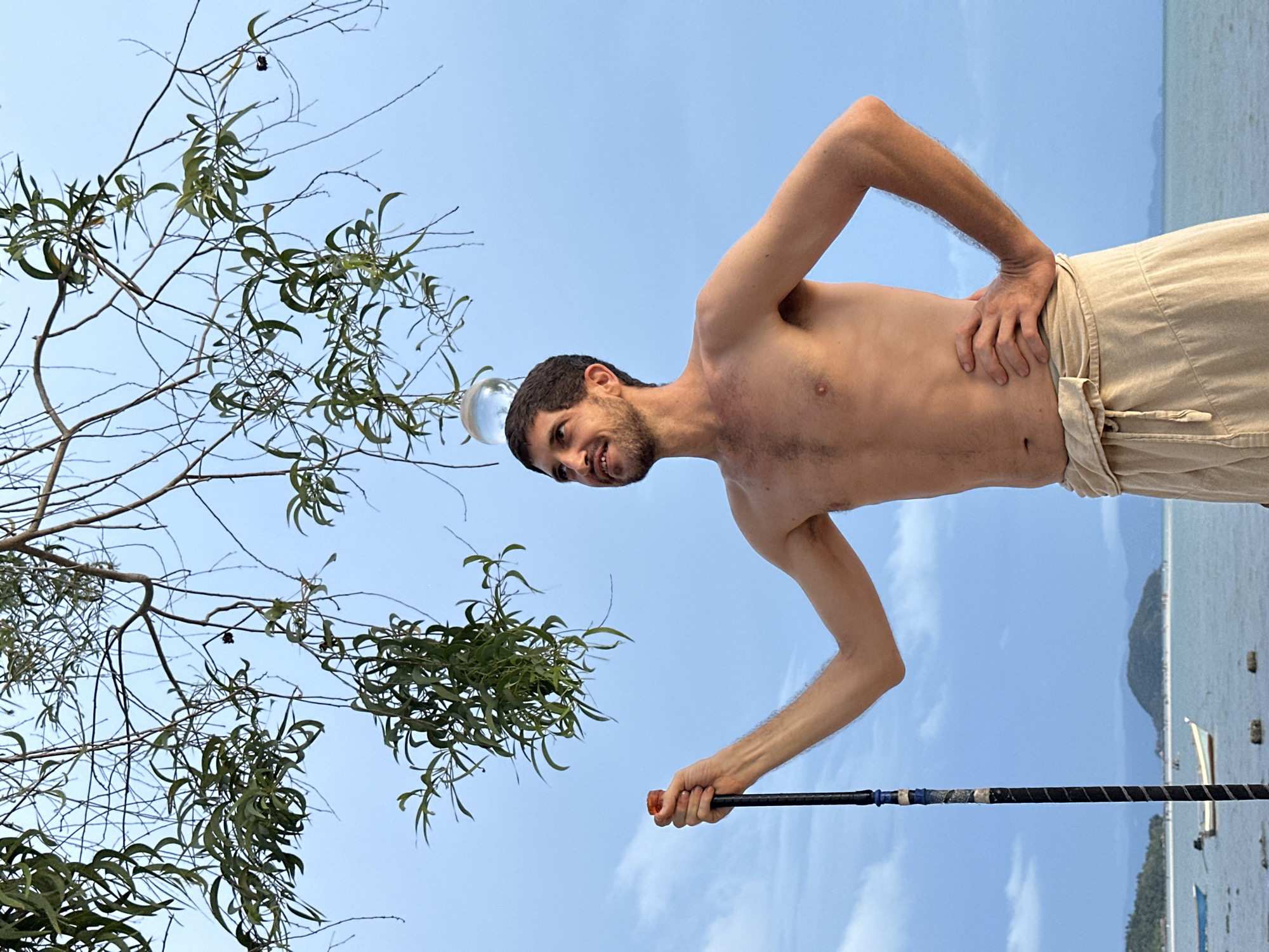 The image shows a shirtless man with a crystal ball balanced on his head, holding a long, black pole. He is wearing tan pants and standing outdoors with a body of water in the background, along with a landmass on the horizon. A tree with slender branches and leaves is to his left. The sky is blue with some clouds. The image was taken in ตำบลเกาะพะงัน, Thailand.