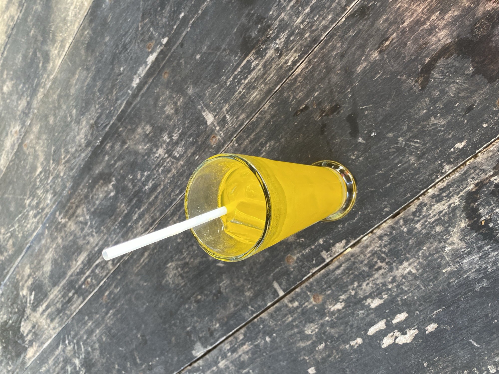 A high-angle, medium shot captures a glass of bright yellow liquid with ice and a white straw, resting on a dark, weathered wooden table. The table's surface is characterized by distinct wood grain and visible wear, with planks running diagonally across the frame. The lighting suggests an outdoor setting, possibly a cafe or restaurant, with dappled sunlight casting shadows and highlights on the table. The image does not depict any people or specific landmarks of Lamkruet, Indonesia, other than the presumed presence of the table and drink within that location.