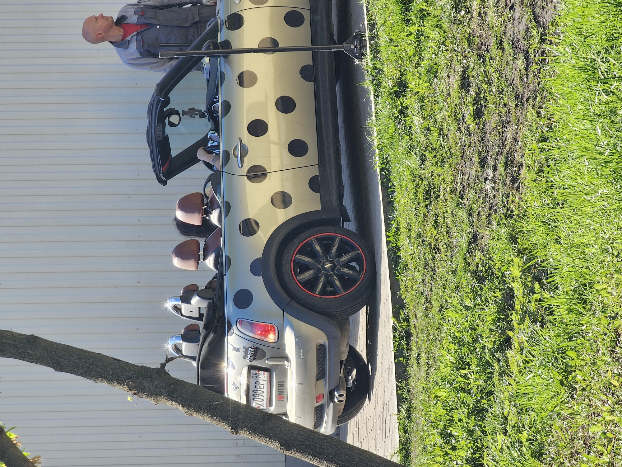 The image shows a modified Mini Cooper convertible parked next to a patch of grass in what appears to be a city. The car has a beige body with large black polka dots. It has four seats, all covered in brown leather, visible in the open-top design. A bald man, wearing a gray and red uniform, is standing near the front of the car, next to an opened door. The license plate reads “X090EP 98 RUS," and it is partially obscured by a sticker. The location is likely Khimki, Russia based on the license plate series. The environment includes a building with light-colored horizontal siding in the background, grass on the side, and a tree branch in the bottom left corner. The car is parked on a paved surface.