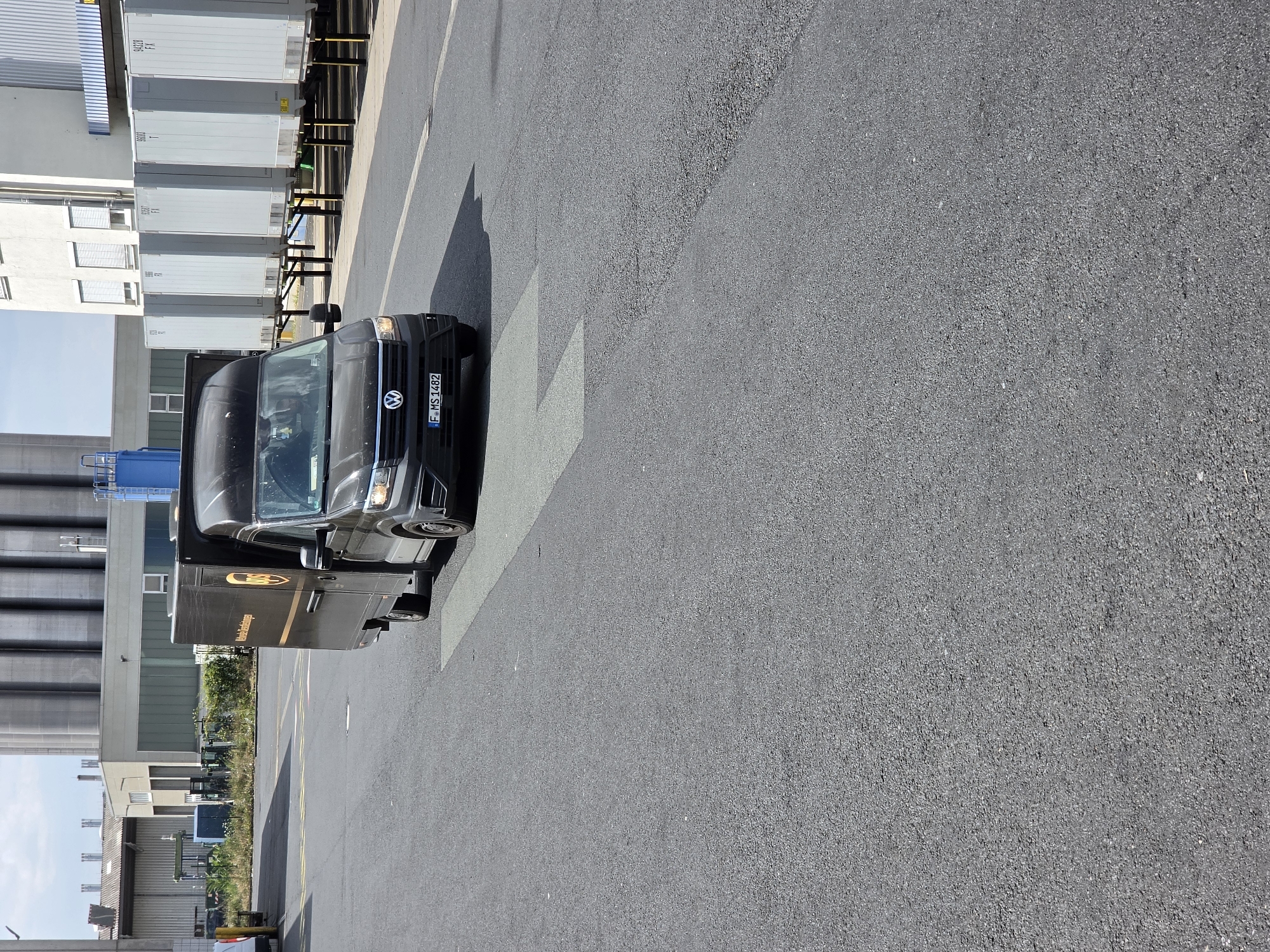 A dark brown UPS delivery van with a license plate reading "F-MS 1482" is driving on an asphalt road. The van has a Volkswagen logo on the front. In the background, there are several parked shipping containers raised on legs, and industrial buildings. The scene appears to be an industrial area or a logistics hub. The weather is clear with a partly cloudy sky.